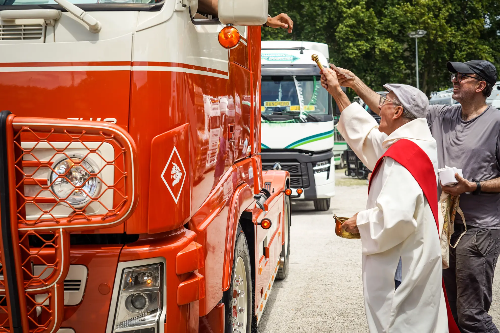 Un sacerdote bendice un camión rojo y a su conductor al aire libre, con otro hombre de pie a su lado.