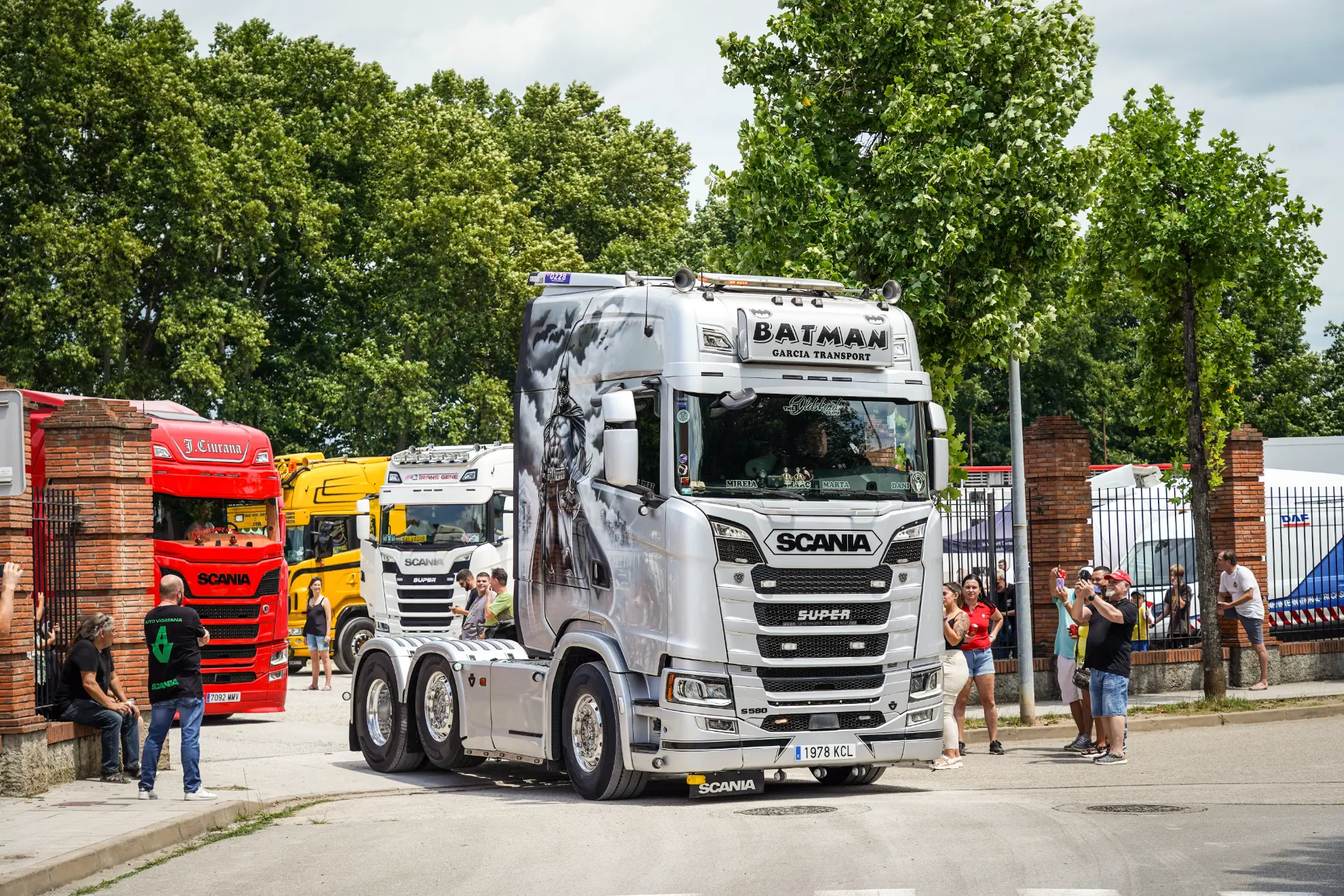 Un camión Scania plateado con ilustraciones de Batman encabeza la lista de otros camiones coloridos mientras la gente toma fotografías al aire libre.