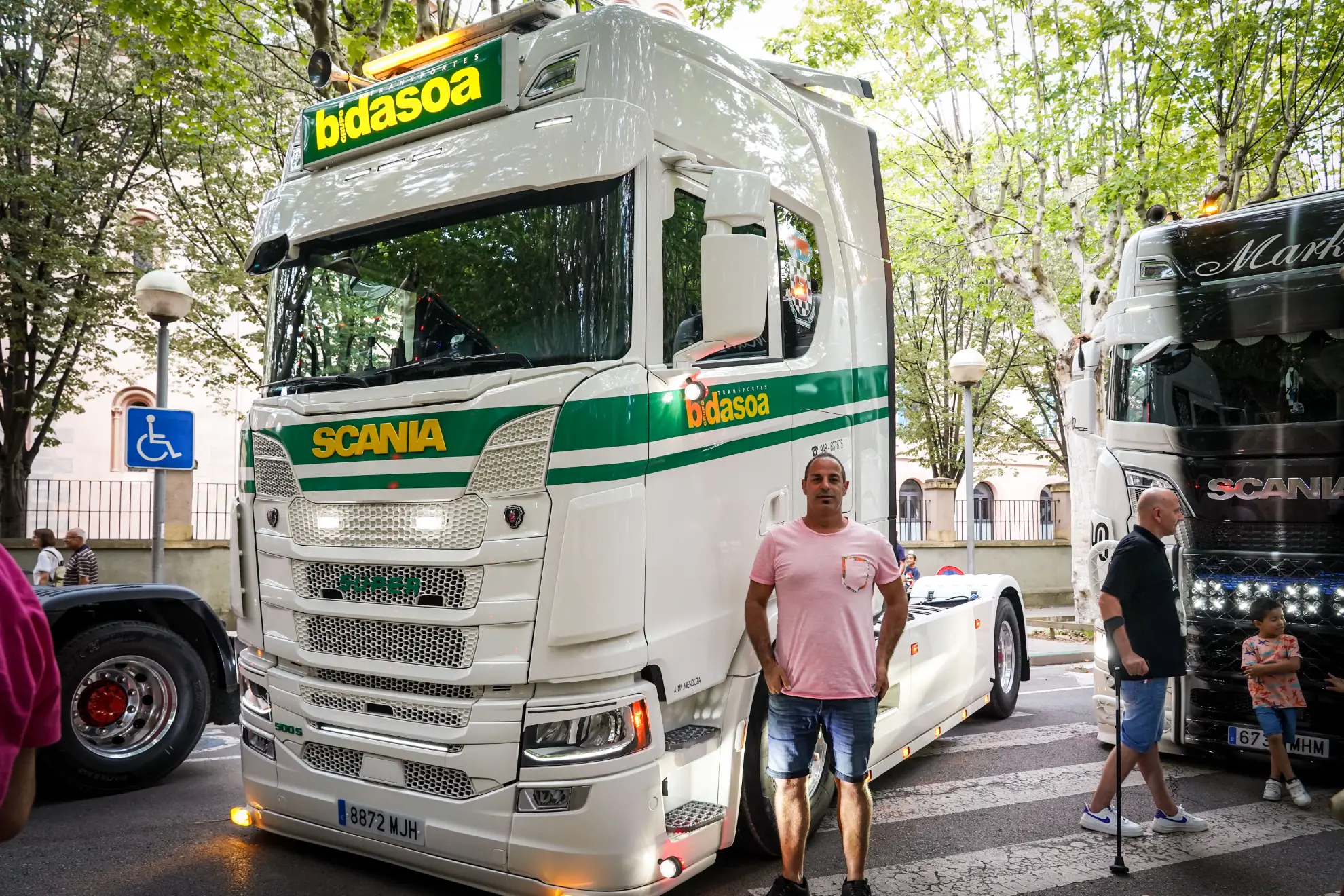 Un hombre con una camisa rosa está parado frente a un camión Scania blanco con la palabra “bidasoa” en la parte superior, estacionado en la calle, como si revisara un borrador automático.