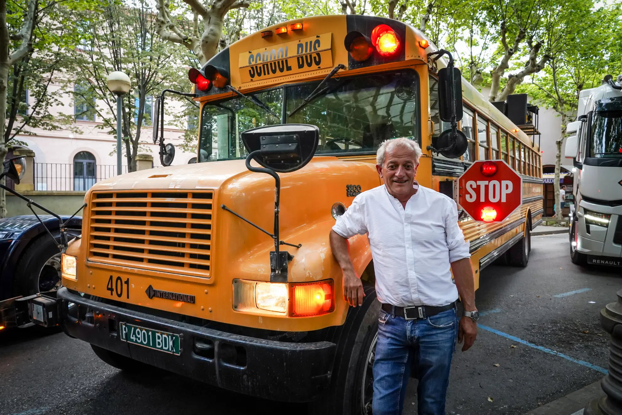 Un hombre sonriente con una camisa blanca se encuentra frente a un autobús escolar amarillo con una señal de pare extendida, y parece como si estuviera listo para comenzar su borrador automático del día.