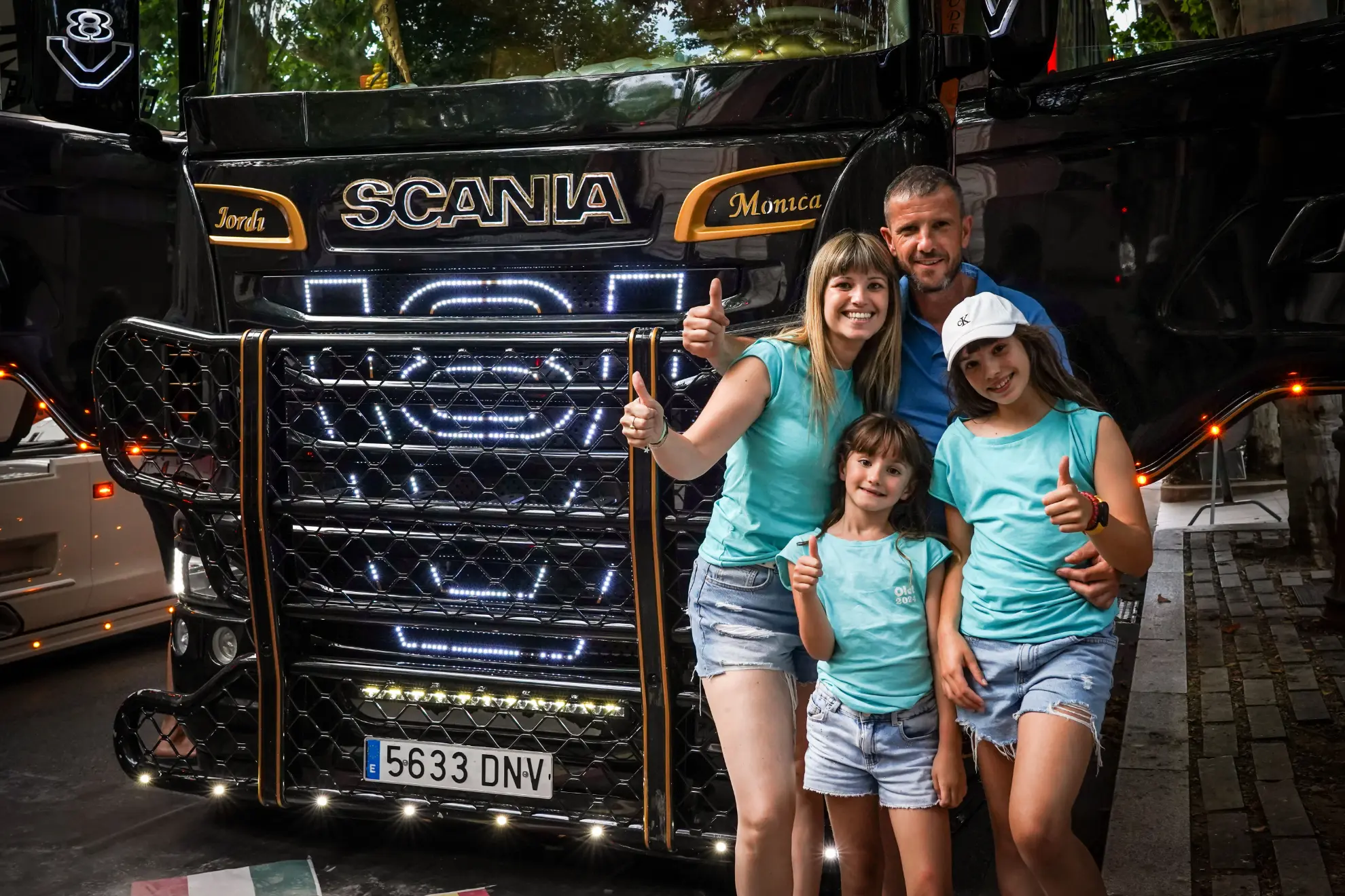 Una familia sonriente de cuatro personas con trajes azules iguales posando con los pulgares hacia arriba frente a un camión Scania negro, celebrando su nuevo borrador automático.