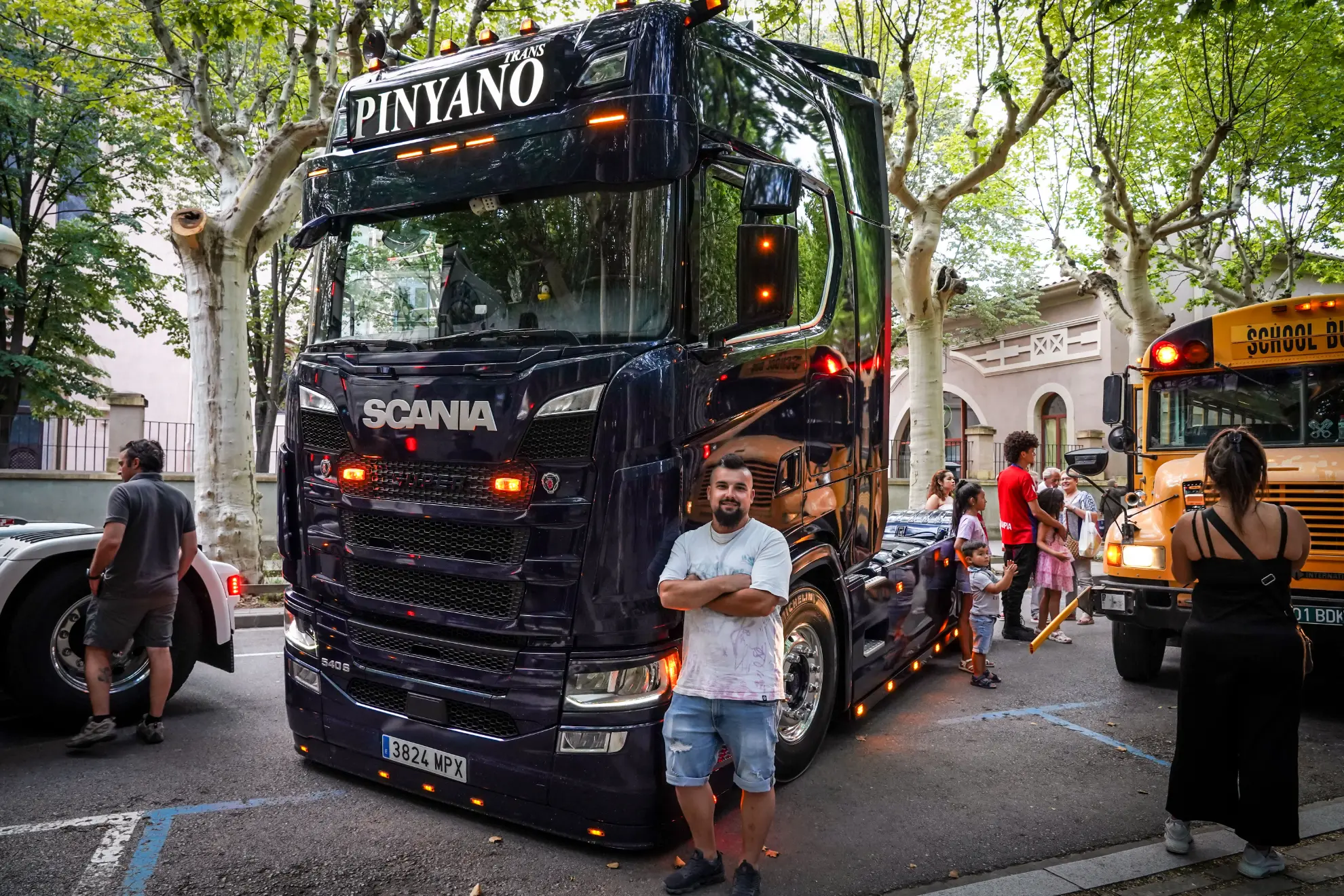 Un hombre está parado frente a un gran camión Scania negro con "PINYANO" en el cartel, estacionado en la calle, al lado de un borrador automático.