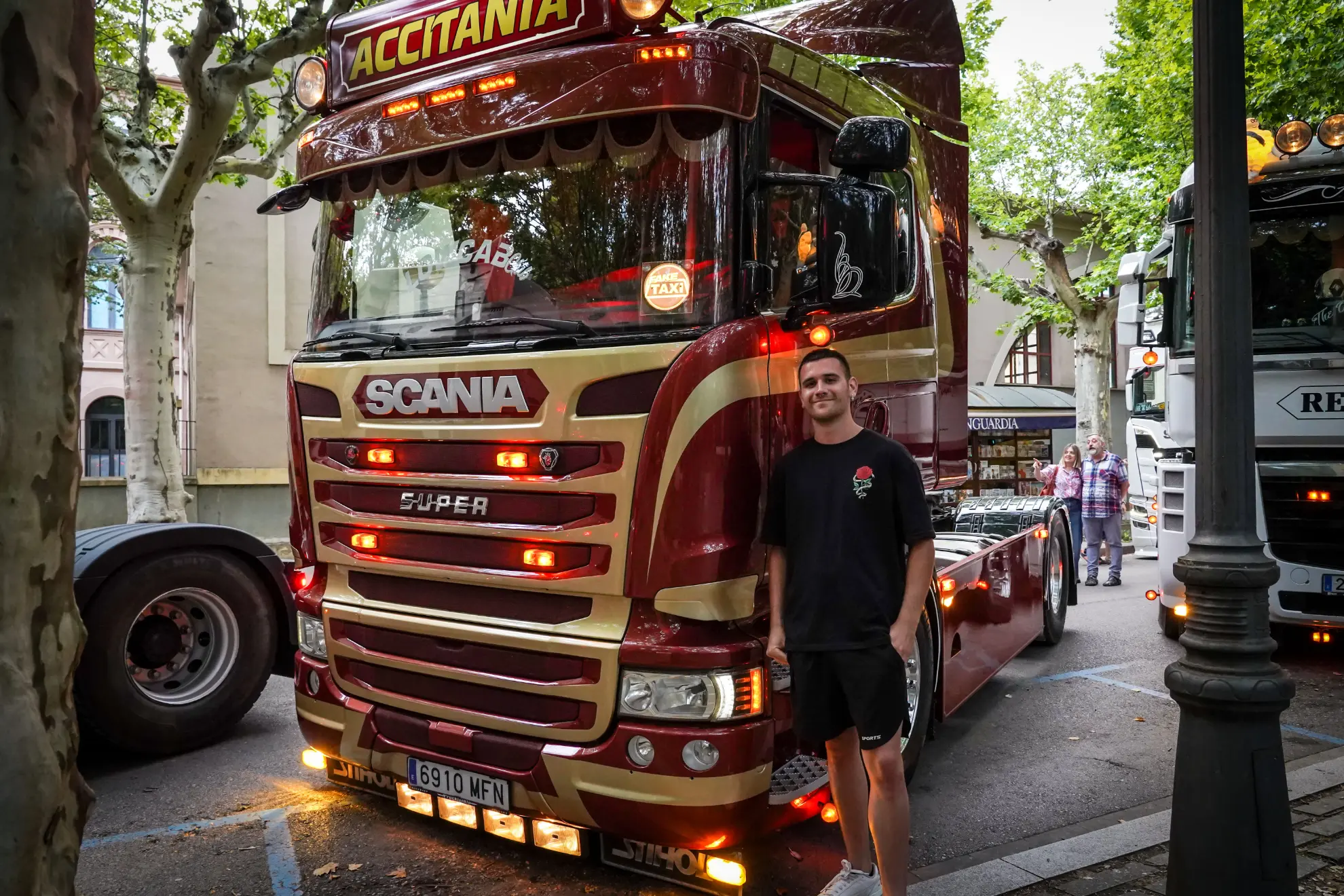 Un hombre con una camisa negra está parado frente a un camión Scania rojo, estacionado en una calle arbolada, revisando algo en su teléfono, tal vez trabajando en un borrador automático.