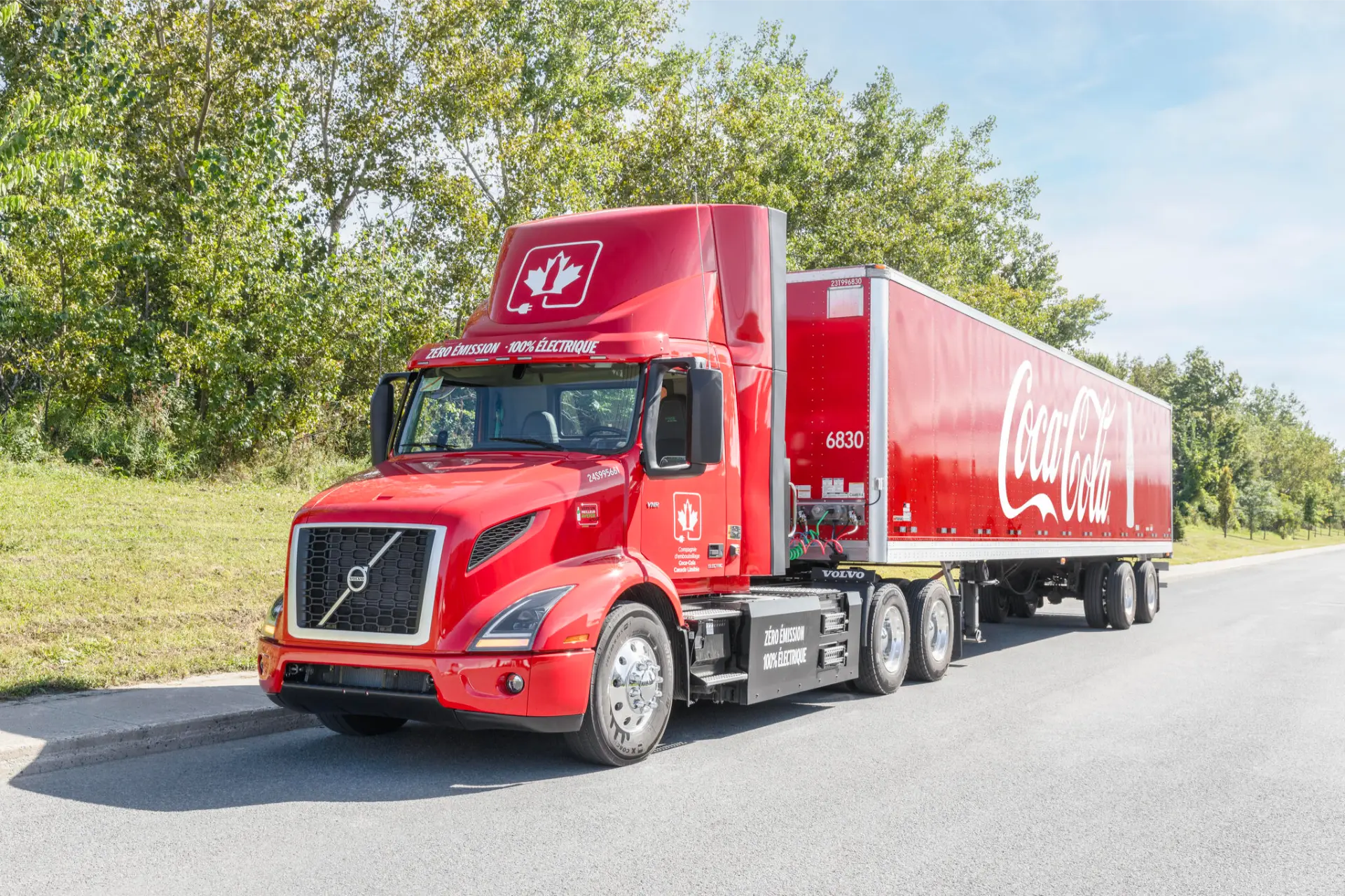 Un camión semirremolque rojo de Coca-Cola, con el logotipo de Borrador automático, está estacionado en una carretera junto a árboles verdes en un día soleado.