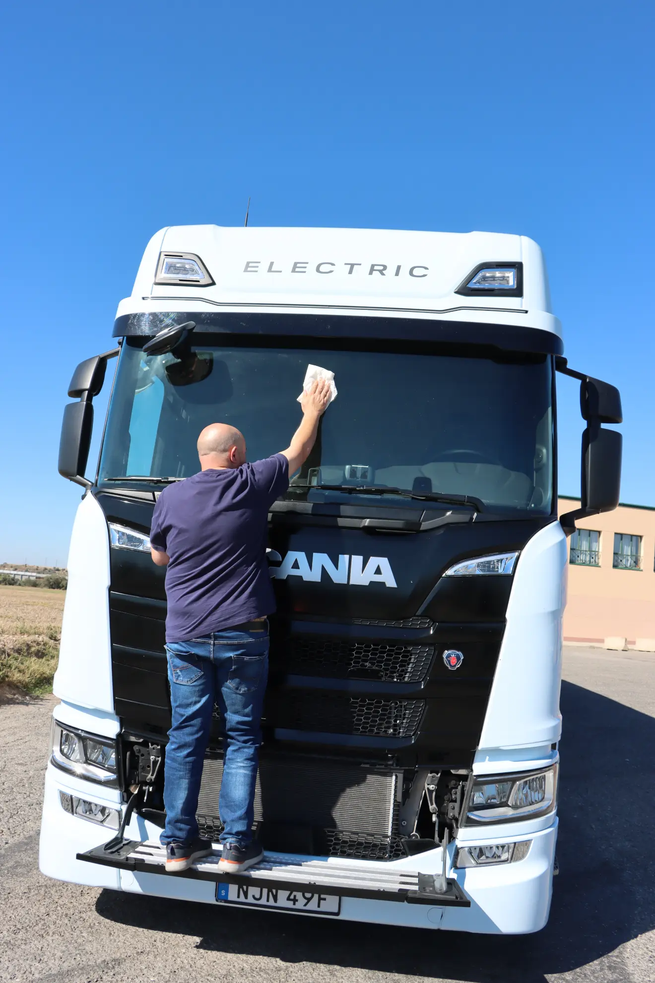 Un hombre limpia el parabrisas de un camión eléctrico Scania 45R blanco estacionado al aire libre bajo un cielo azul claro, preparándolo para una prueba de cambio a eléctrico.