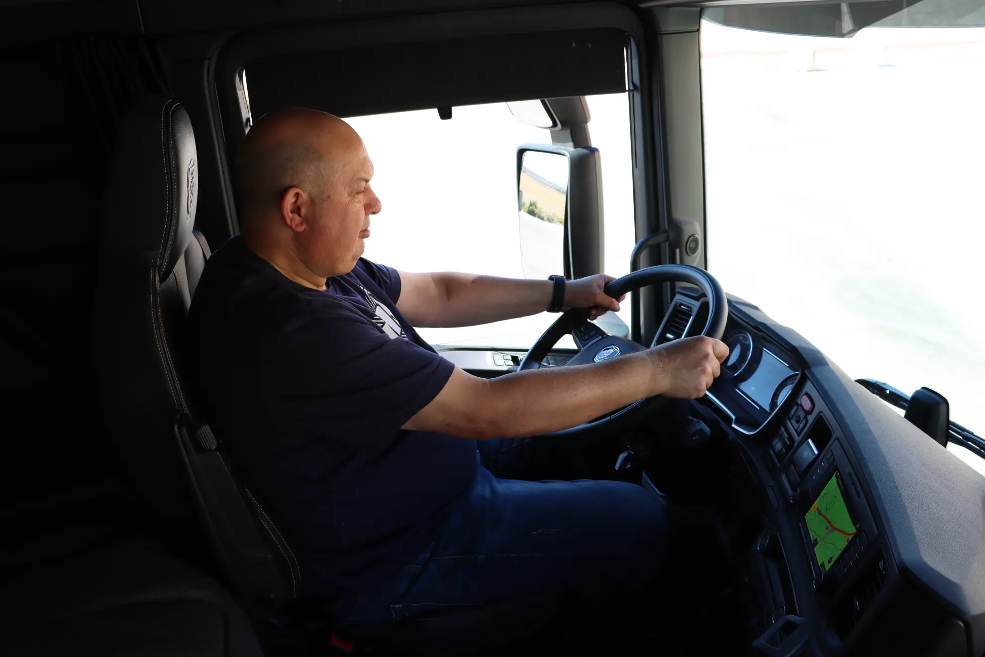 Hombre calvo con camisa oscura conduce un camión Scania 45R, sujetando el volante con ambas manos durante una prueba de cambio a eléctrico.