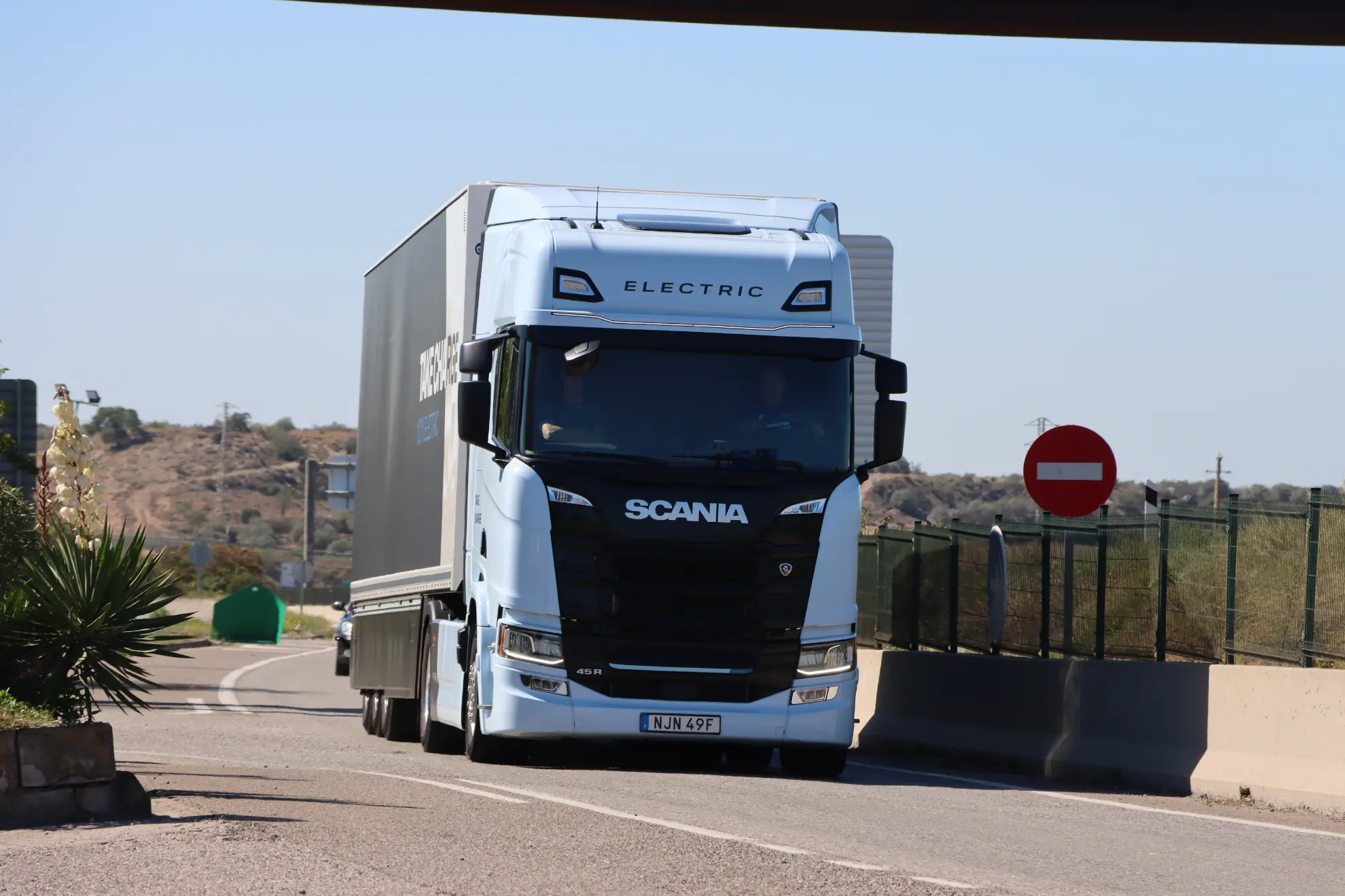 Un camión eléctrico Scania 45R blanco circula por una carretera cerca de una zona cercada y una señal de prohibido el paso, mostrando el cambio a eléctrico durante una prueba.