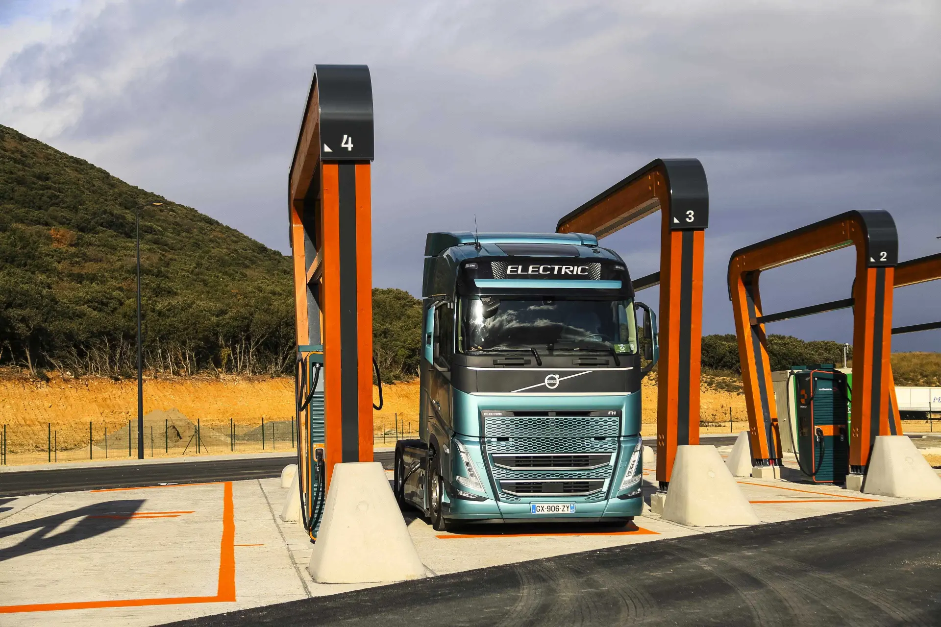 Un camión eléctrico reposa en una estación de carga bajo un cielo nublado, con colinas al fondo, como si estuviera estacionado junto a un borrador automático.