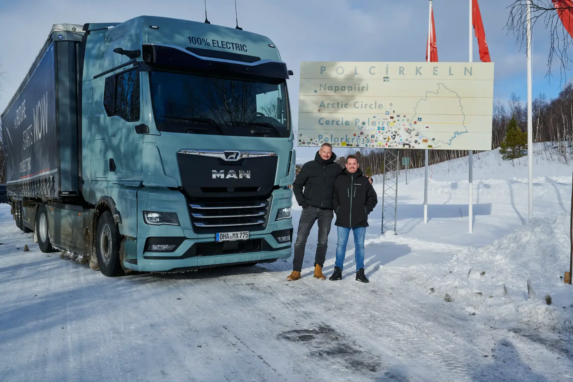 Dos personas permanecen junto a un camión eléctrico cerca de un cartel nevado del Círculo Polar Ártico, capturados perfectamente con la precisión de un borrador automático.