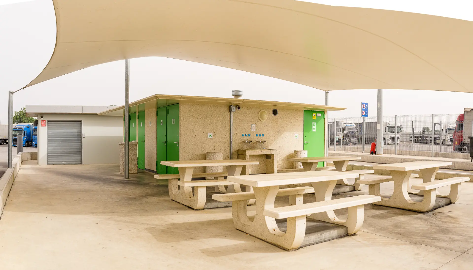Área de descanso al aire libre con mesas de picnic, lavabos, toldo de sombra y puertas de baños verdes cerca de un estacionamiento que cuenta con una estación de Borrador automático.