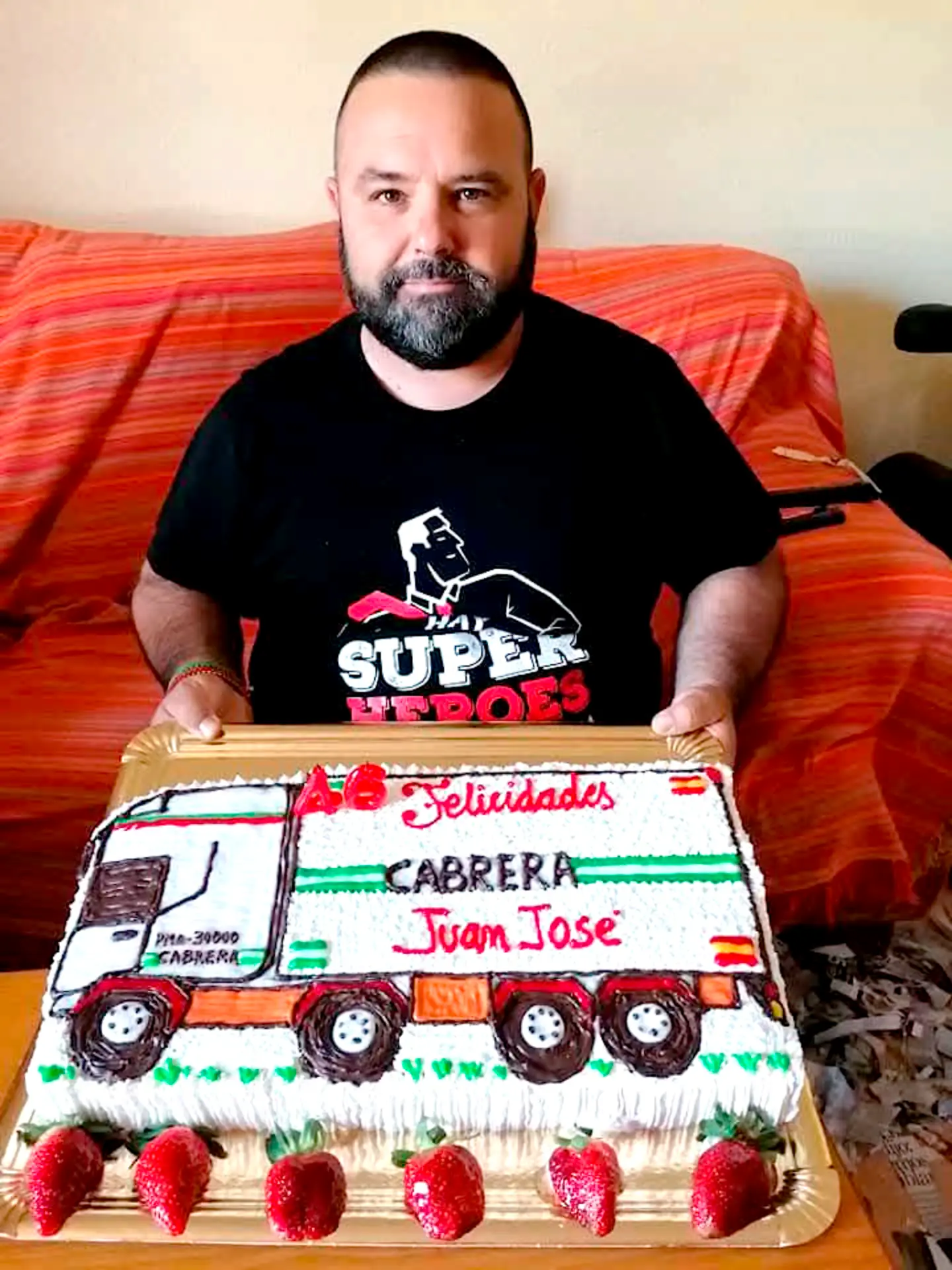 Un hombre con barba sostiene un pastel decorado con un camión colorido y fresas, perfecto para cualquier fanático de las maquetas de camiones.