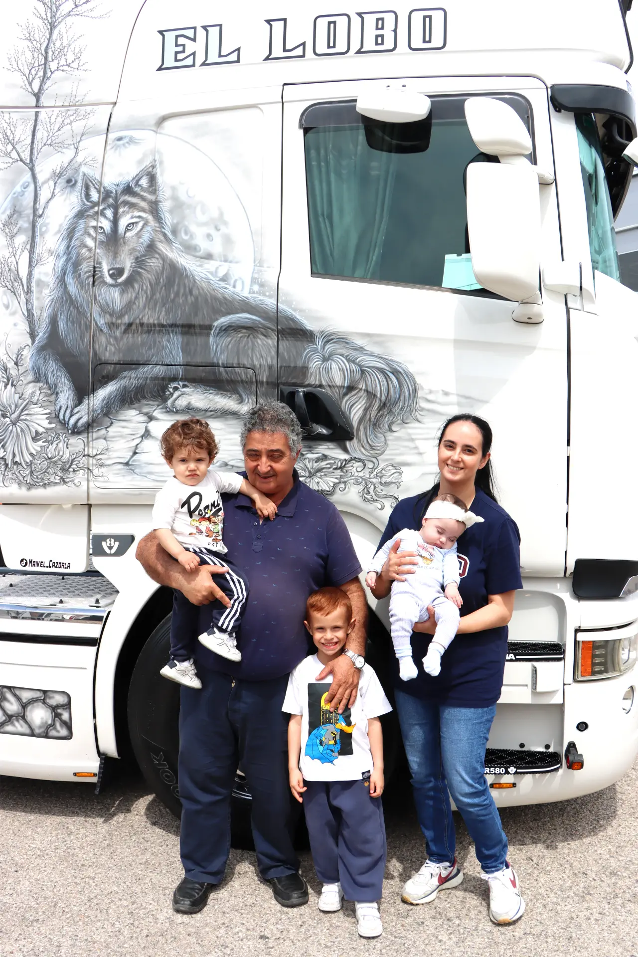 Una familia de cinco posa frente a un camión blanco con un mural de lobo y "EL LOBO" escrito encima en el Valencia Nascar Fest 2025, celebrando la pasión por los camiones americanos tuneados.