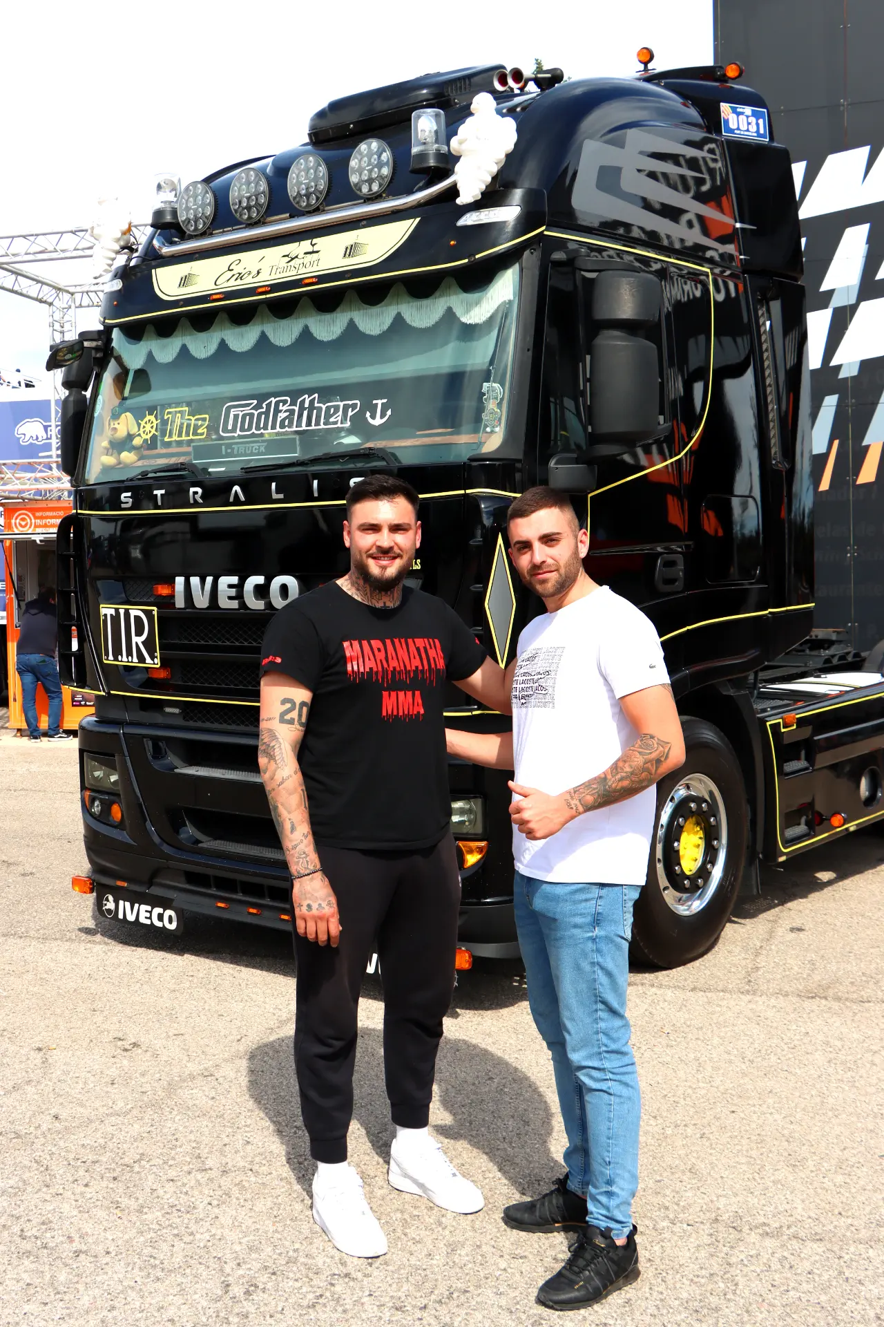 Dos hombres posan sonrientes frente a un gran camión Iveco negro etiquetado como "El Padrino" en un evento al aire libre con camiones americanos tuneados, probablemente parte de la vibrante escena de carreras camiones España.