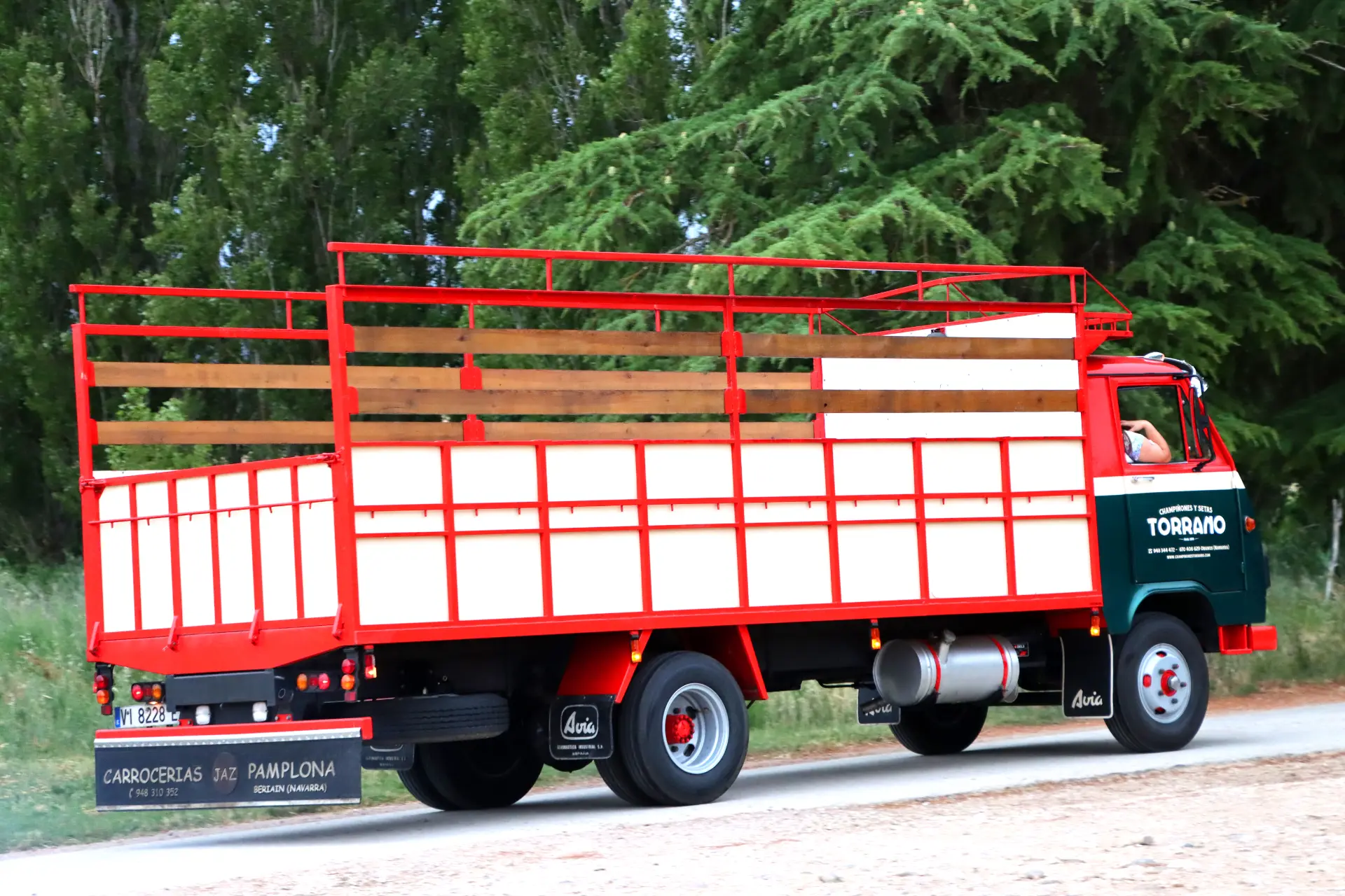 Un camión Avia 5000 rojo y blanco, de plataforma y con rieles de madera, conocido como camión familiar, circula por un camino rural bordeado de árboles verdes.