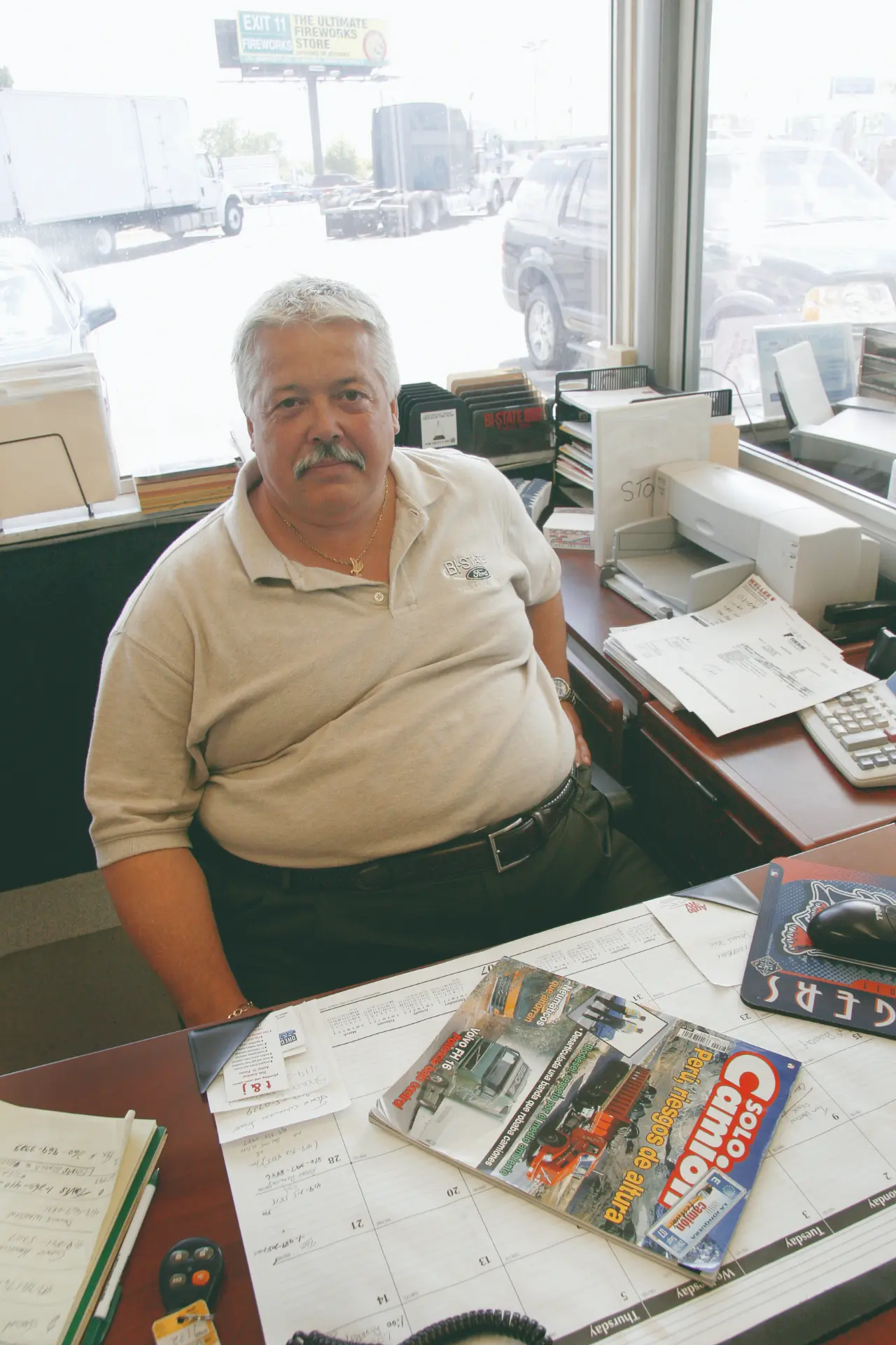 Hombre de pelo gris y bigote sentado en un escritorio con revistas y papeles en una oficina, revisando informes sobre inspecciones en carreteras y controles a camiones en EE.UU.