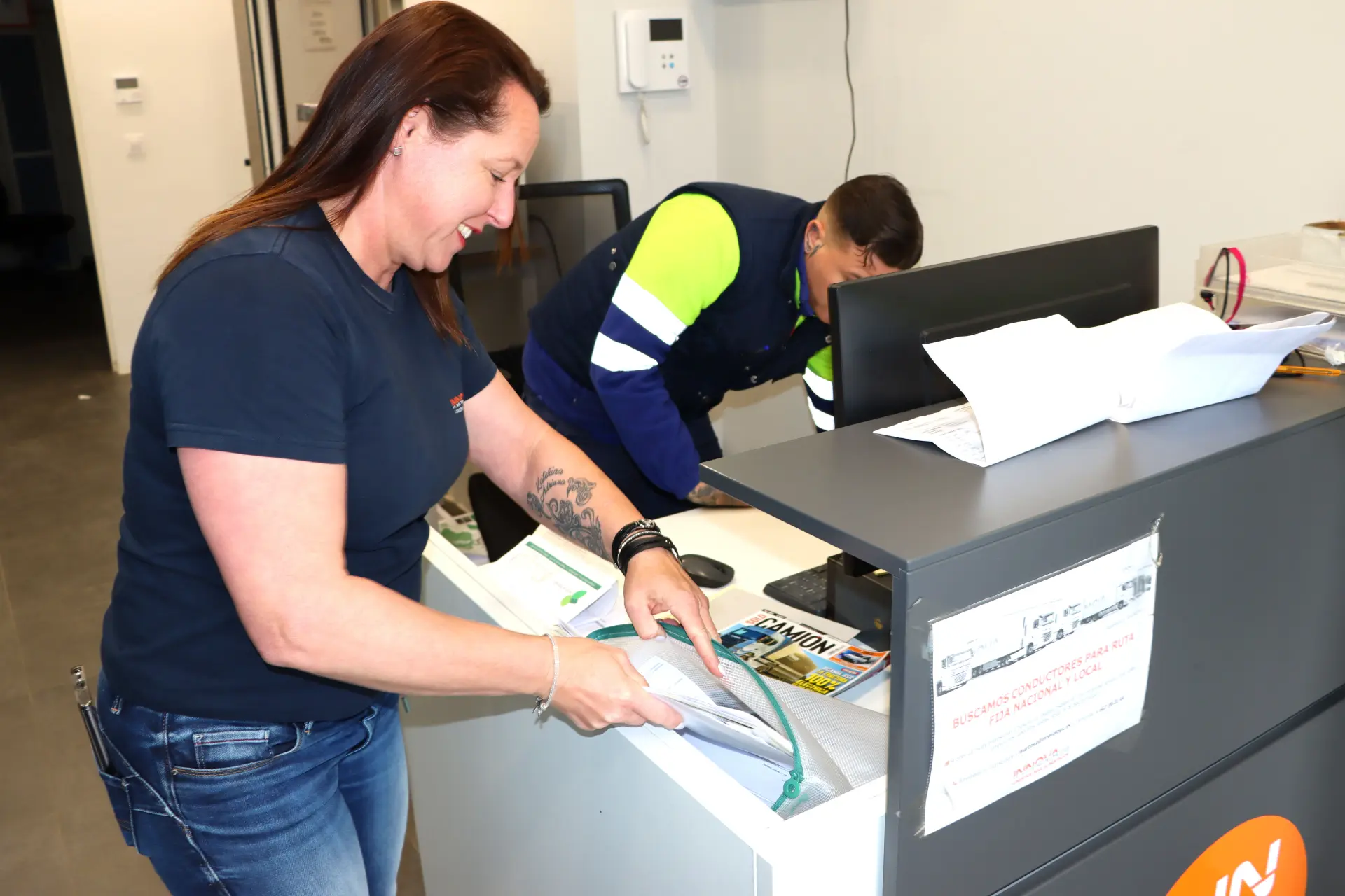 Mujer sonriendo mientras organiza papeles en un mostrador de recepción, posiblemente preparando un borrado automático; hombre trabajando en una computadora en el fondo.