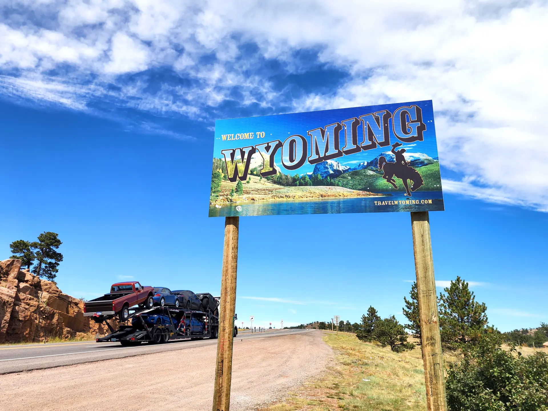 Letrero de “Bienvenido a Wyoming” junto a una carretera bajo un cielo azul, con un camión transportador de automóviles al fondo, que resalta los Controles de camiones y las inspecciones comunes en la carretera.
