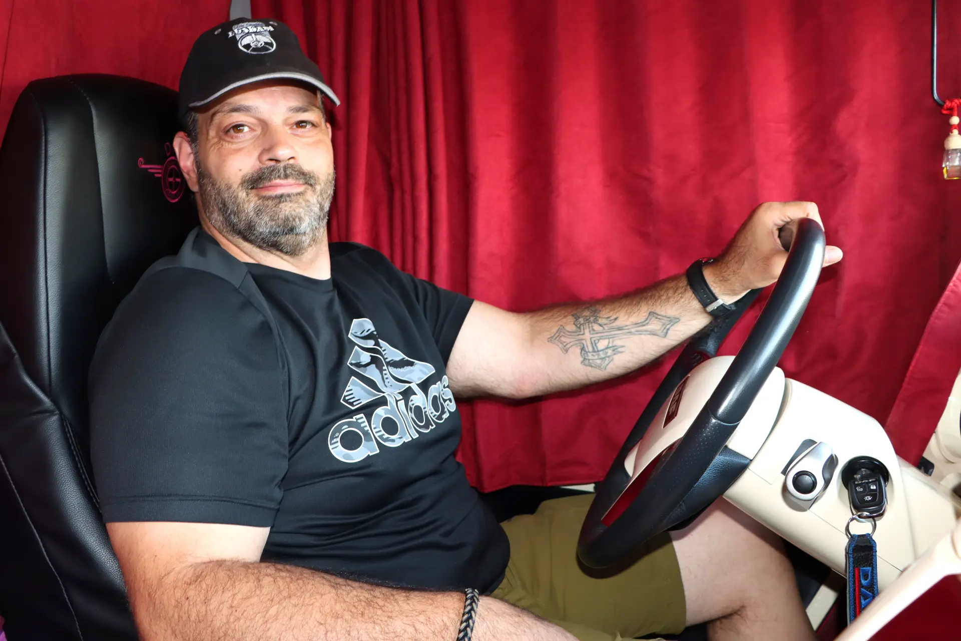 Un hombre con barba se sienta en el asiento del conductor de un camión, sonriendo, vistiendo una gorra, una camiseta Adidas negra y pantalones cortos.