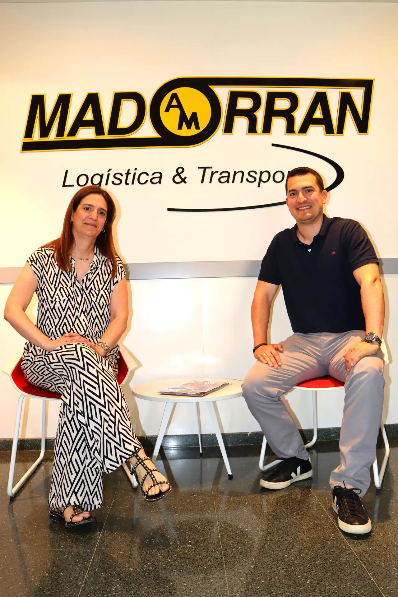 Dos personas están sentadas en sillas frente a un cartel de "Madorran Logística & Transport" en la pared, revisando un borrador automático.