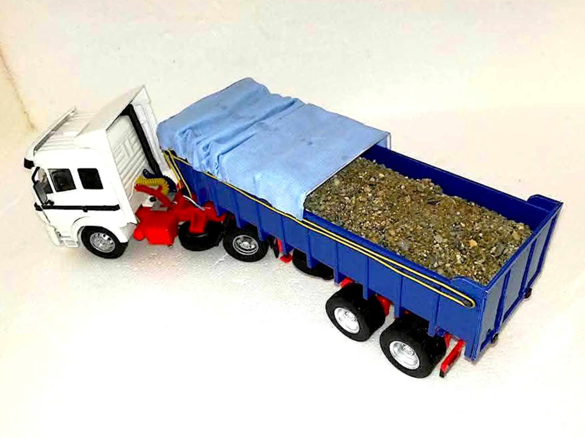 Un camión volquete de juguete a escala 1:24 con un remolque azul, parcialmente cubierto y lleno de grava o tierra, perfecto para maquetas de camiones, exhibido sobre un fondo blanco.