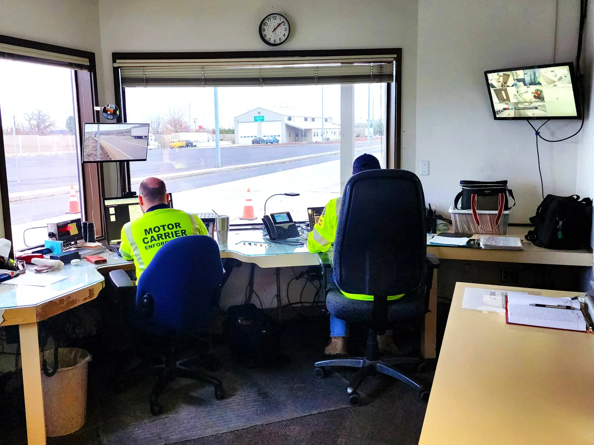 Dos trabajadores con chalecos de alta visibilidad se sientan en escritorios en una oficina con grandes ventanas y monitores de vigilancia, revisando un borrador automático en sus pantallas.