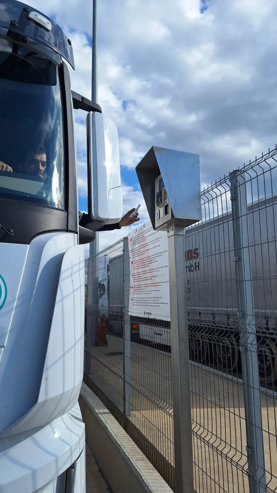 Un camionero usa un borrador automático para alcanzar por la ventana una caja de metal en un puesto de control vallado bajo un cielo nublado.