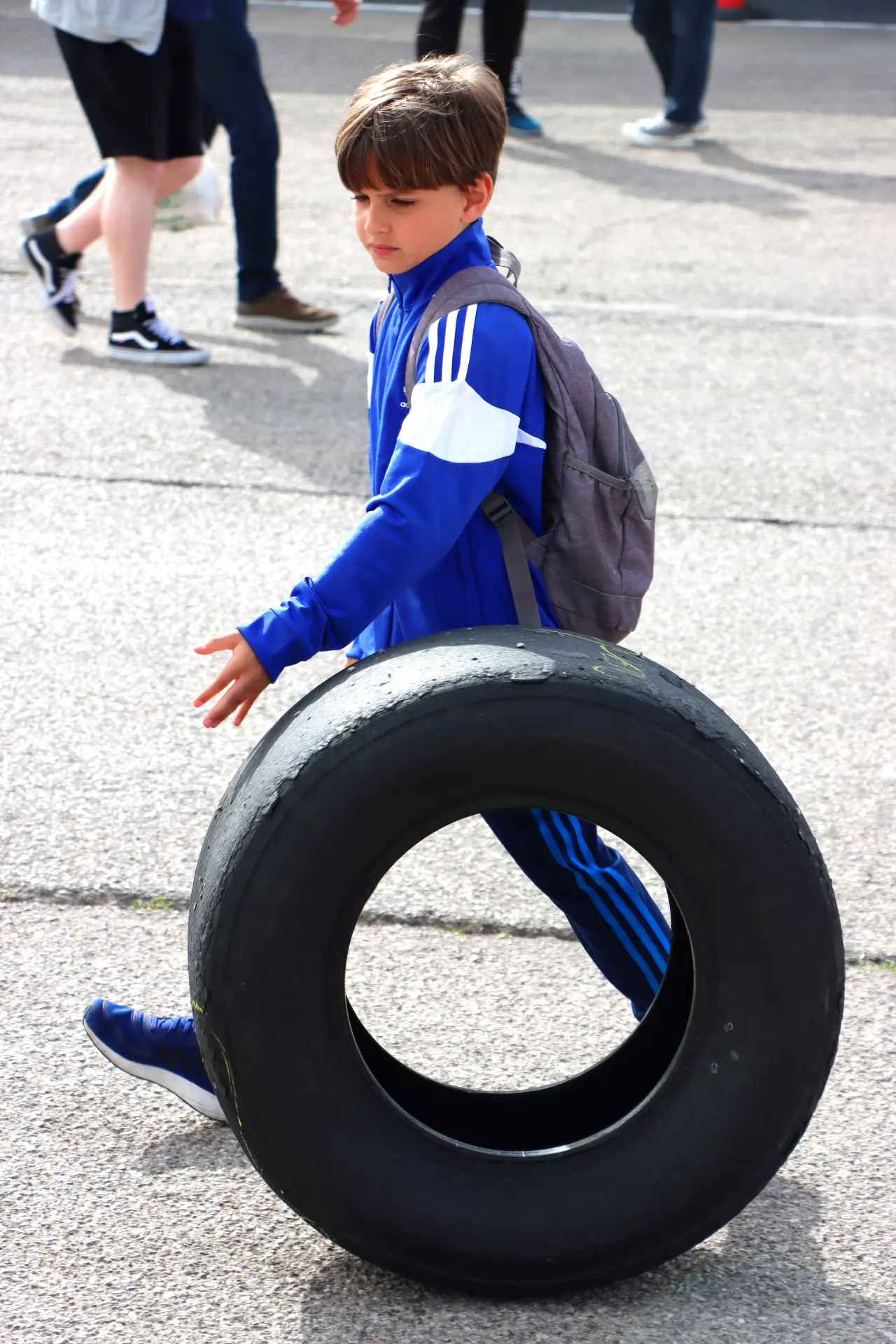 Un niño con un chándal azul hace rodar un neumático grande sobre una superficie pavimentada al aire libre, inspirado en la emoción de las carreras de camiones España y la emoción del Valencia NASCAR Fest 2025.