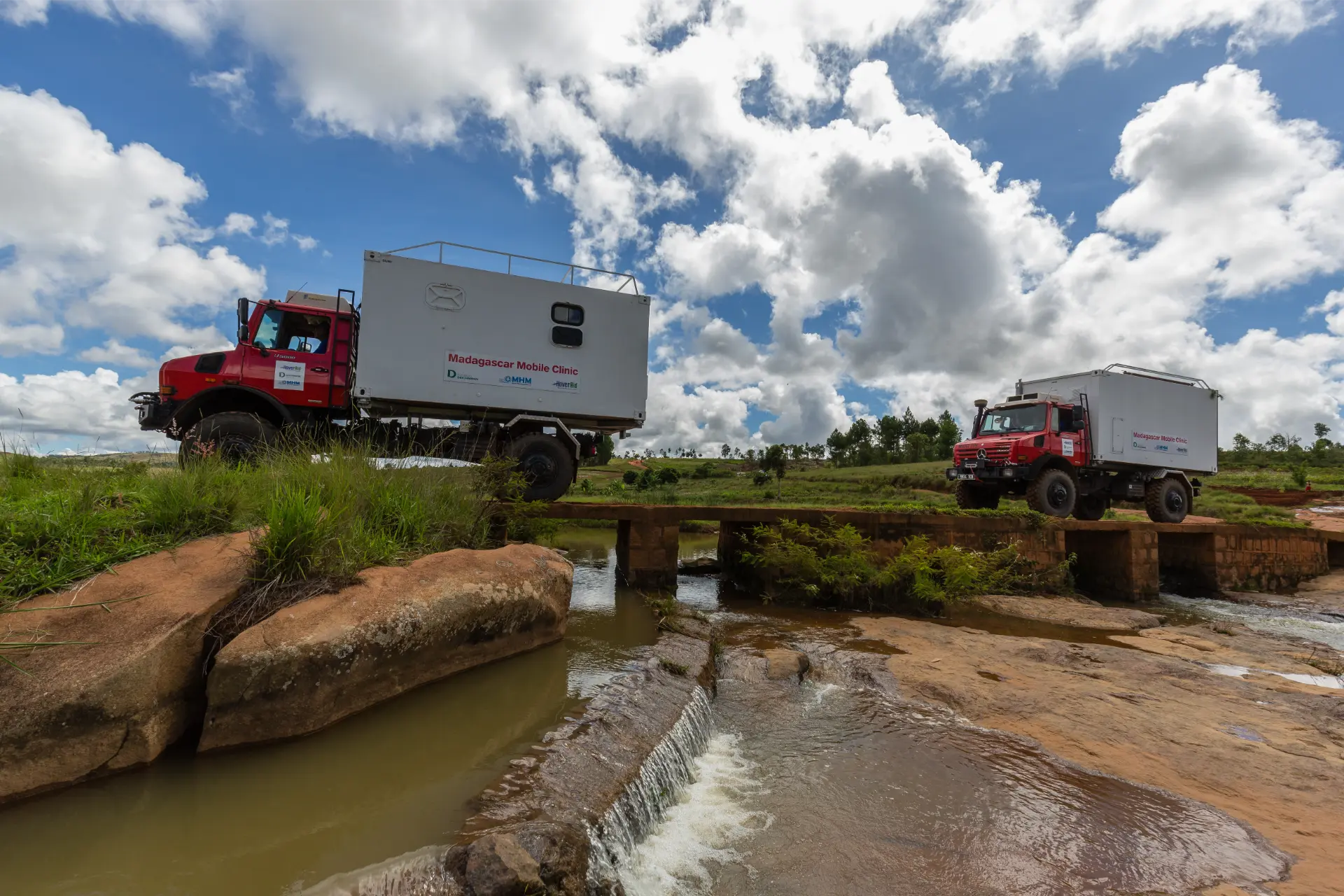 Dos camiones rojos de una clínica móvil cruzan un puente estrecho sobre un pequeño arroyo, con terreno cubierto de hierba y cielo nublado.