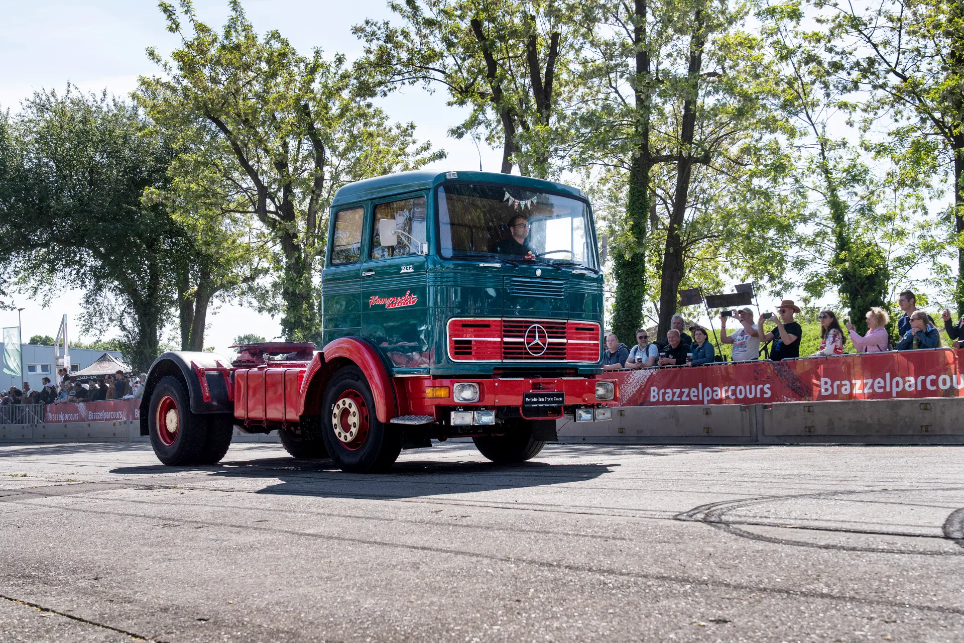 Un camión Mercedes-Benz antiguo verde y rojo, resaltado por sus detalles Borrador automático, pasa junto a una multitud en un evento al aire libre.