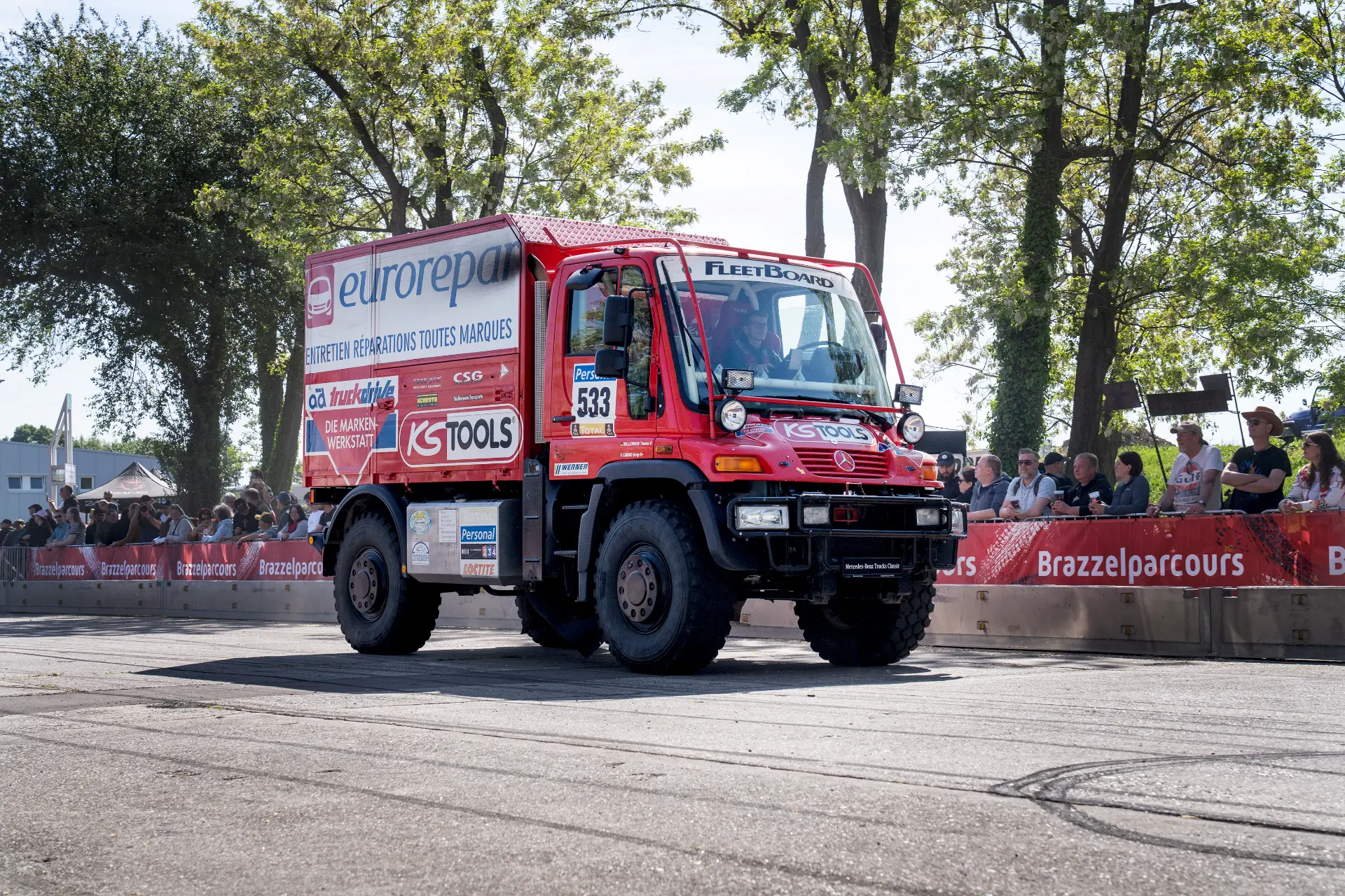 Un camión de rally rojo equipado con un borrador automático corre en un circuito callejero bordeado de espectadores y árboles en un día soleado.