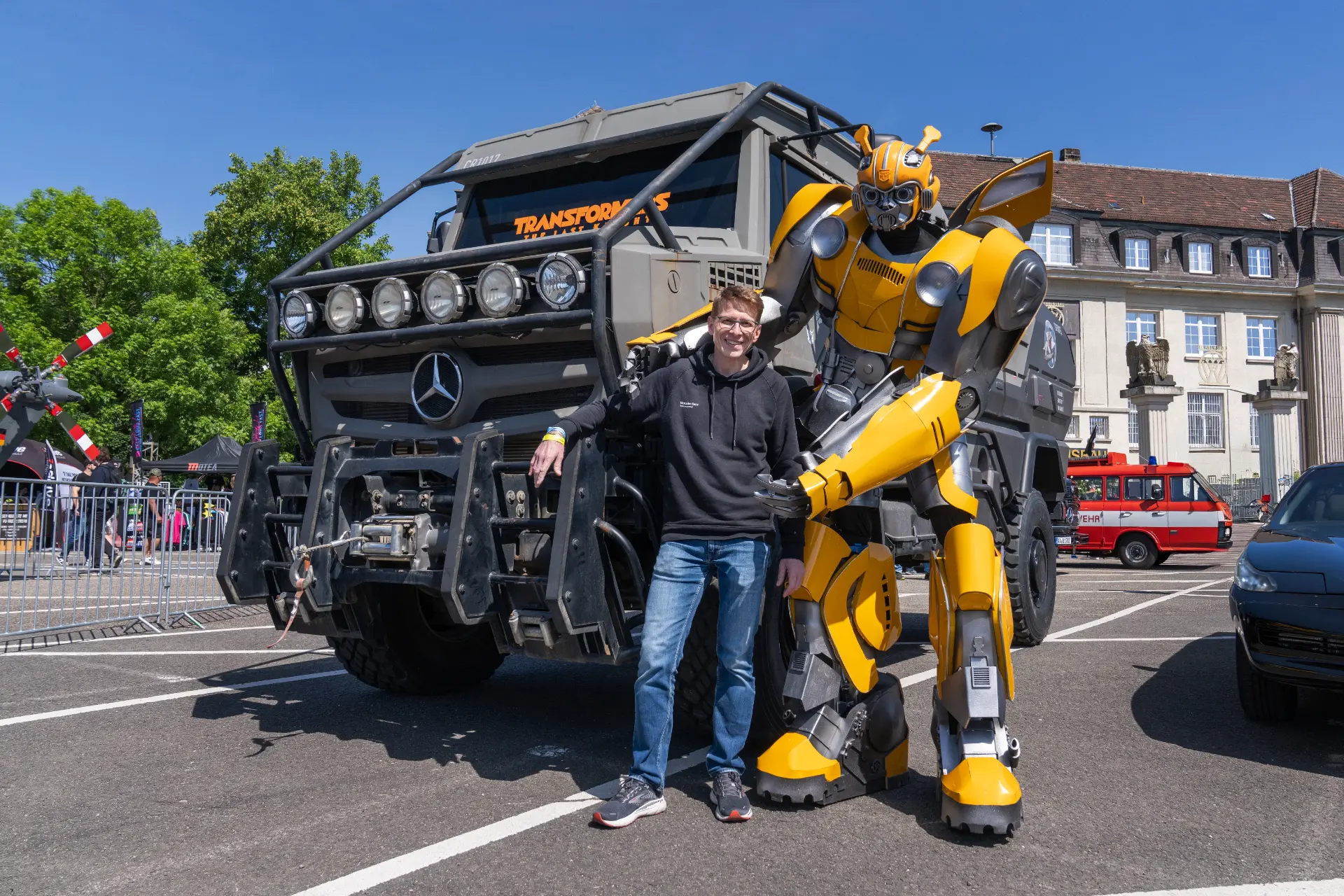 Un hombre posa con un robot Bumblebee junto a un gran camión Mercedes en un evento al aire libre, mostrando la impresionante tecnología del Borrador automático.
