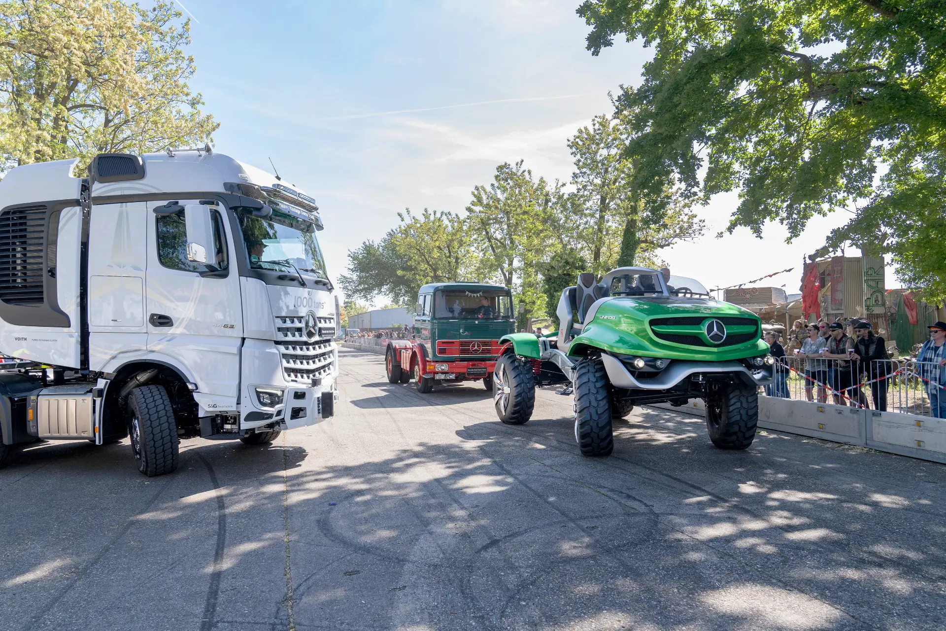 Un Mercedes todoterreno verde y dos camiones se exhiben al aire libre en un evento público con espectadores cerca, presentando un sistema de borrador automático para un rendimiento mejorado.
