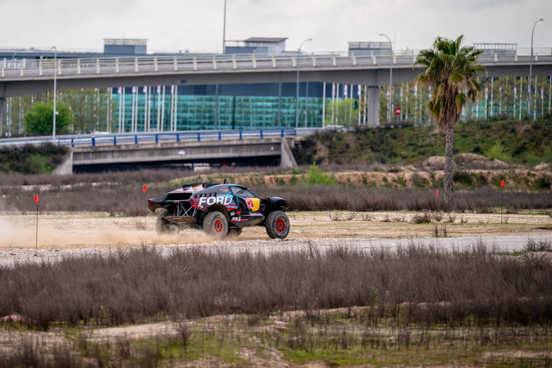 Un coche de rally Ford Performance F1 acelera sobre una pista de tierra, levantando polvo cerca de un puente y una palmera, evocando la emoción del Gran Premio de Madrid 2026.