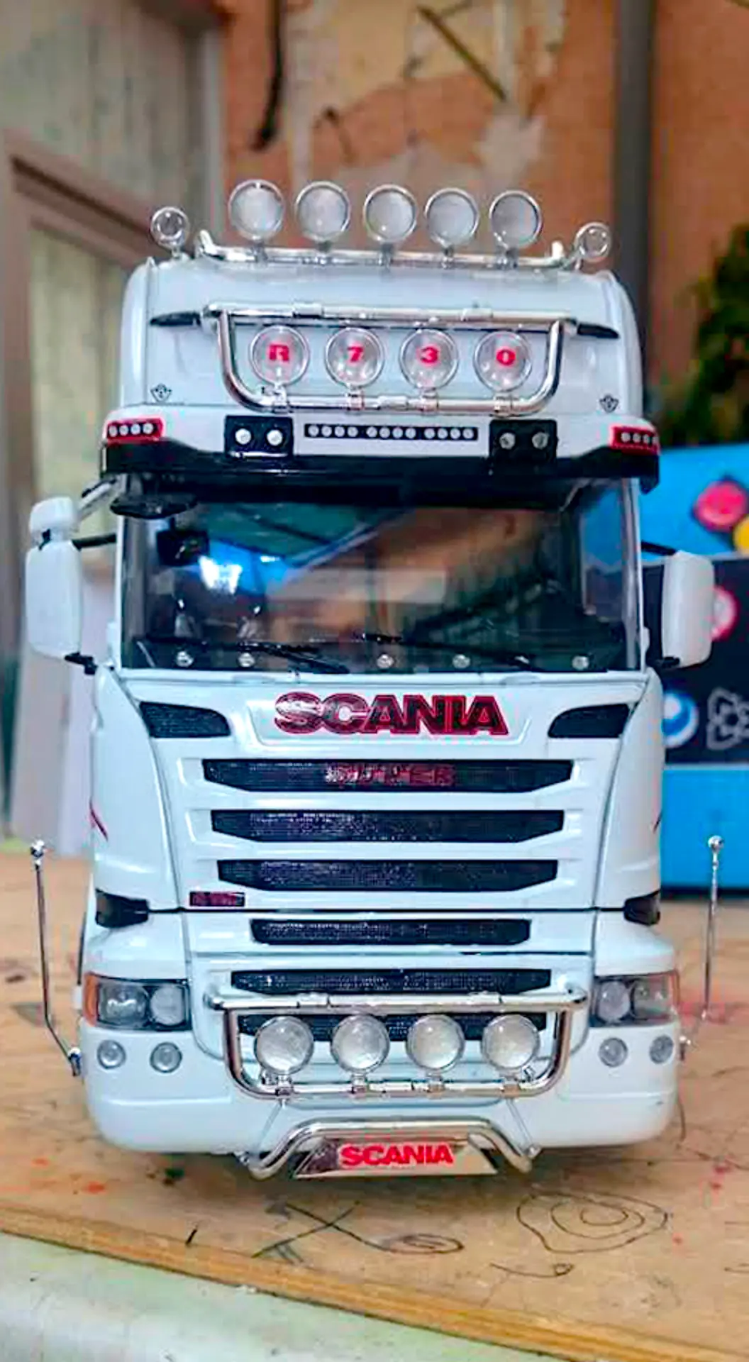 Vista frontal de un camión Scania blanco a escala 1:24 con múltiples luces y detalles en rojo, estacionado en el interior, perfecto para los fanáticos de las maquetas de camiones y cualquier camionero apasionado.