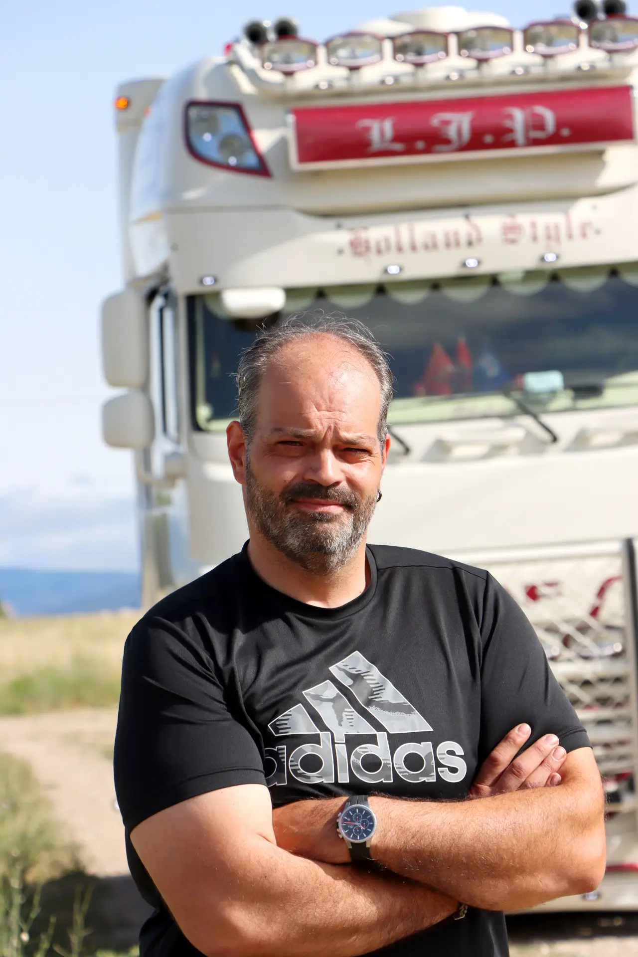 Un hombre con los brazos cruzados frente a un camión semirremolque blanco en un día soleado, vistiendo una camiseta Adidas negra, resalta la eficiencia moderna del borrador automático en los flujos de trabajo de transporte.