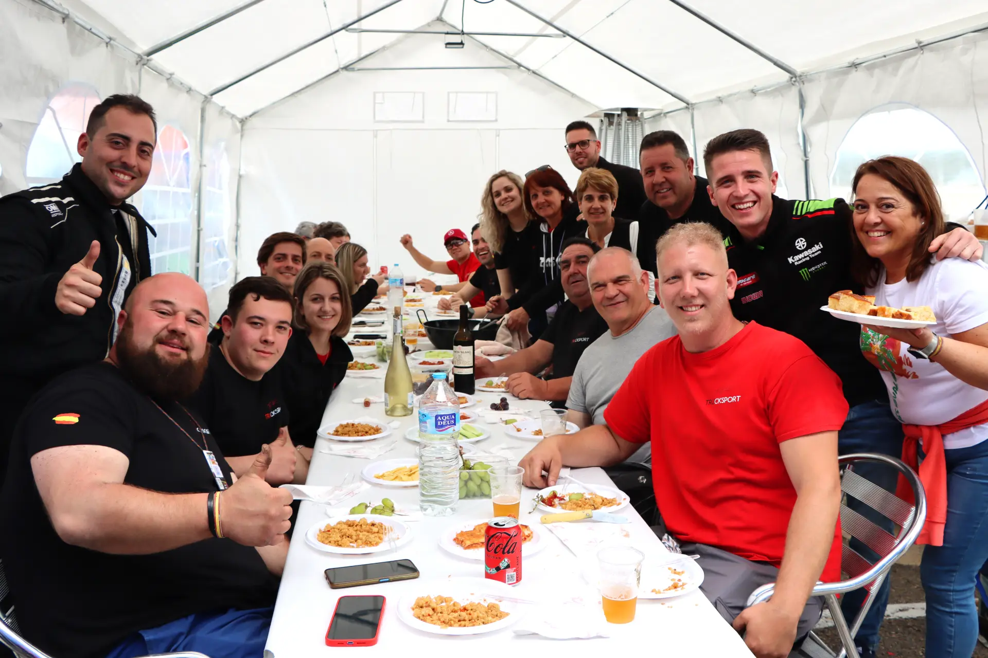 Un grupo grande de personas se sienta alrededor de una mesa larga bajo una carpa en el Valencia NASCAR Fest 2025, sonriendo y disfrutando de una comida juntos mientras camiones americanos tuneados se alinean en el fondo.