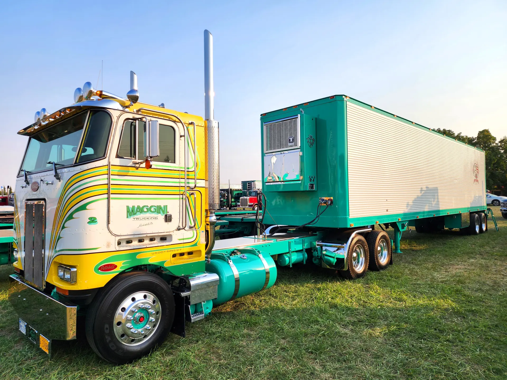 Un camión semirremolque antiguo verde y amarillo con un remolque a juego, rotulado "Borrador automático", está estacionado en el césped bajo un cielo despejado.