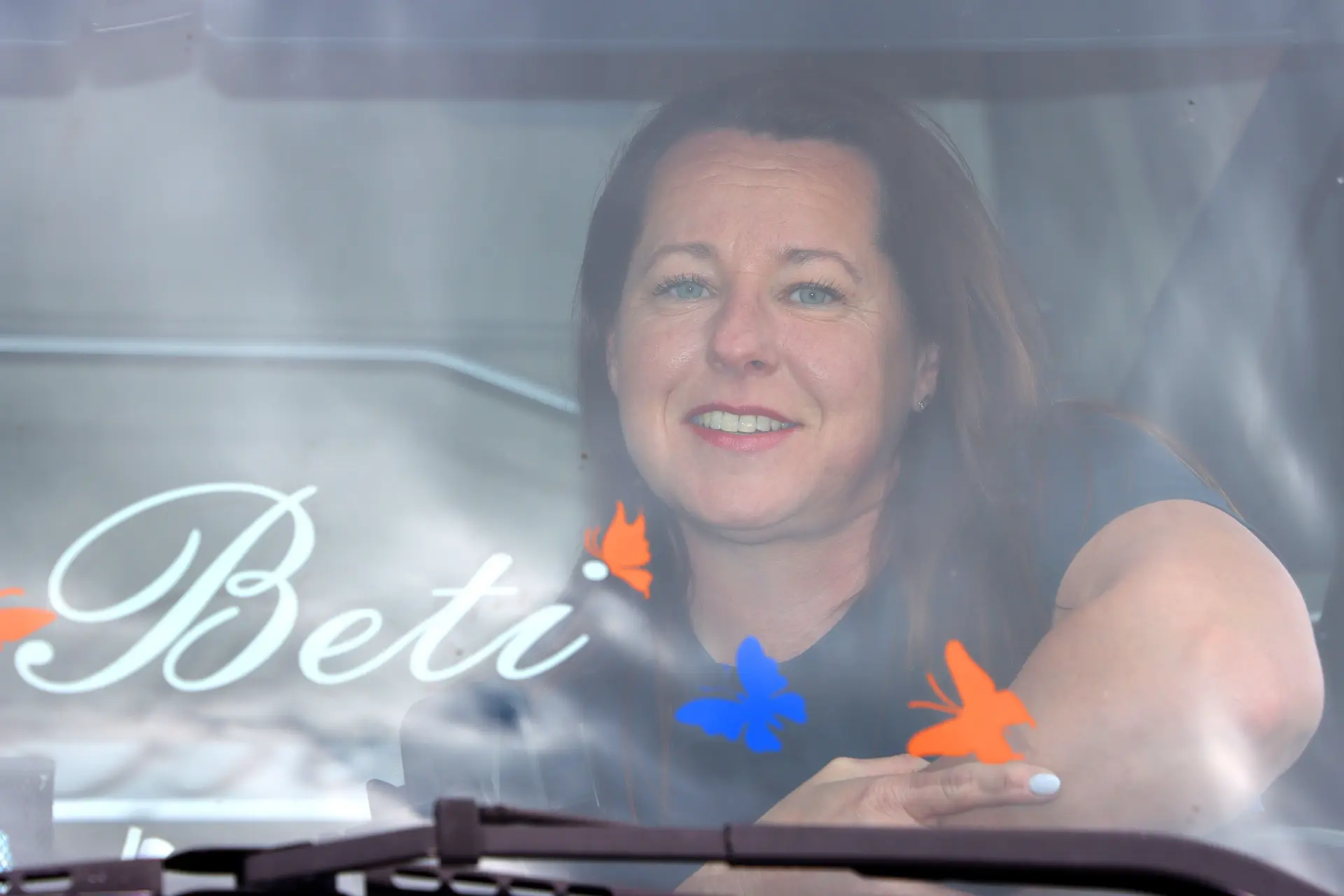 Una mujer sonriente se sienta detrás de un parabrisas decorado con mariposas y la palabra “Beti”, como si celebrara los momentos de la vida con un toque de magia de borrador automático.