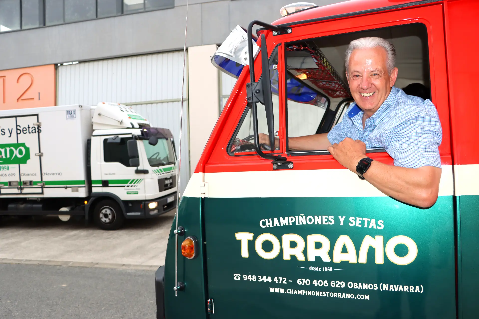 Un hombre sonriente con una camisa azul se sienta en una camioneta TORRANO roja y verde, equipada con borrador automático, con un camión de reparto en el fondo.