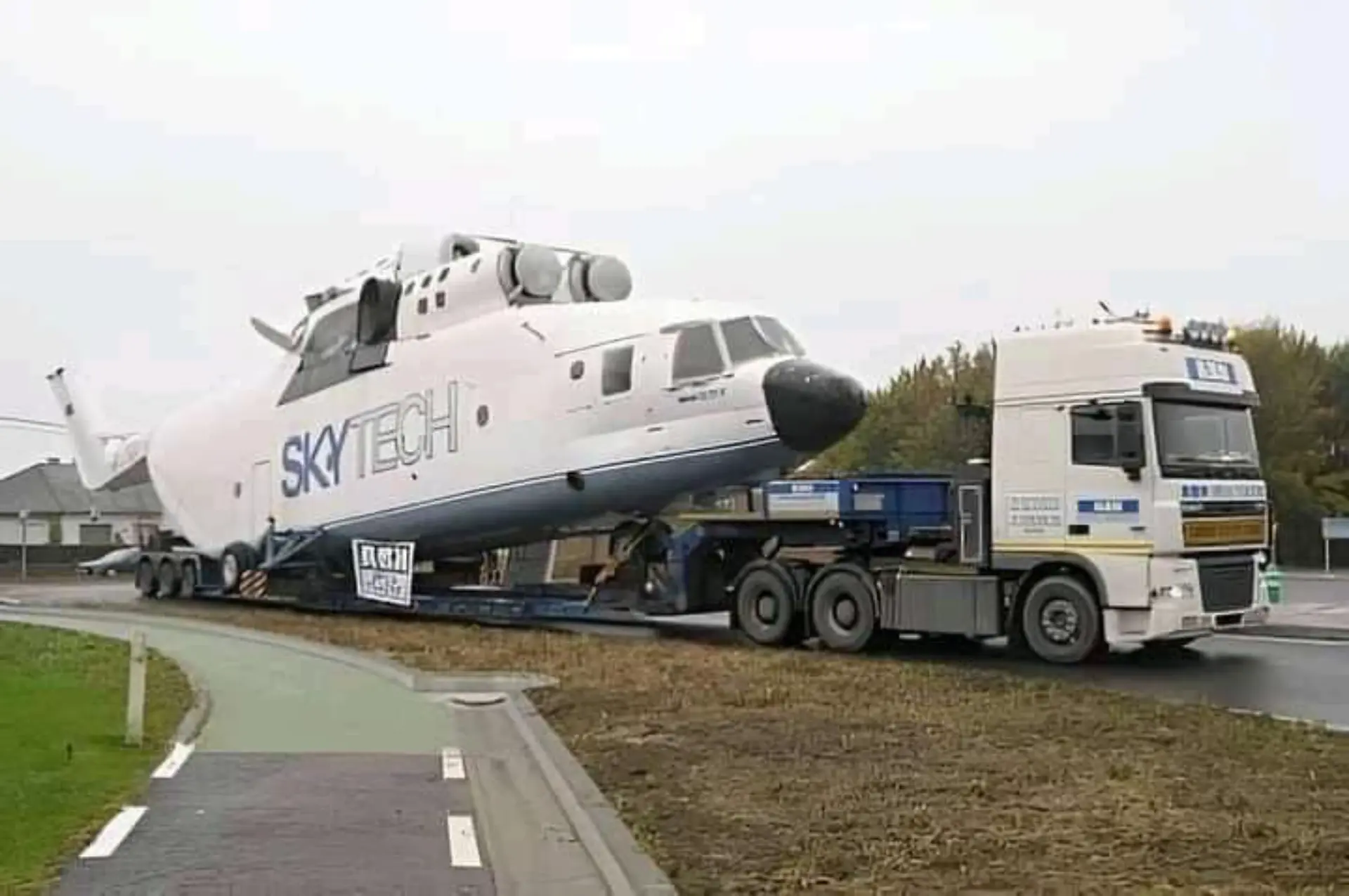 Un camión grande transporta el fuselaje de un helicóptero SkyTech blanco en un remolque de plataforma por una carretera.
