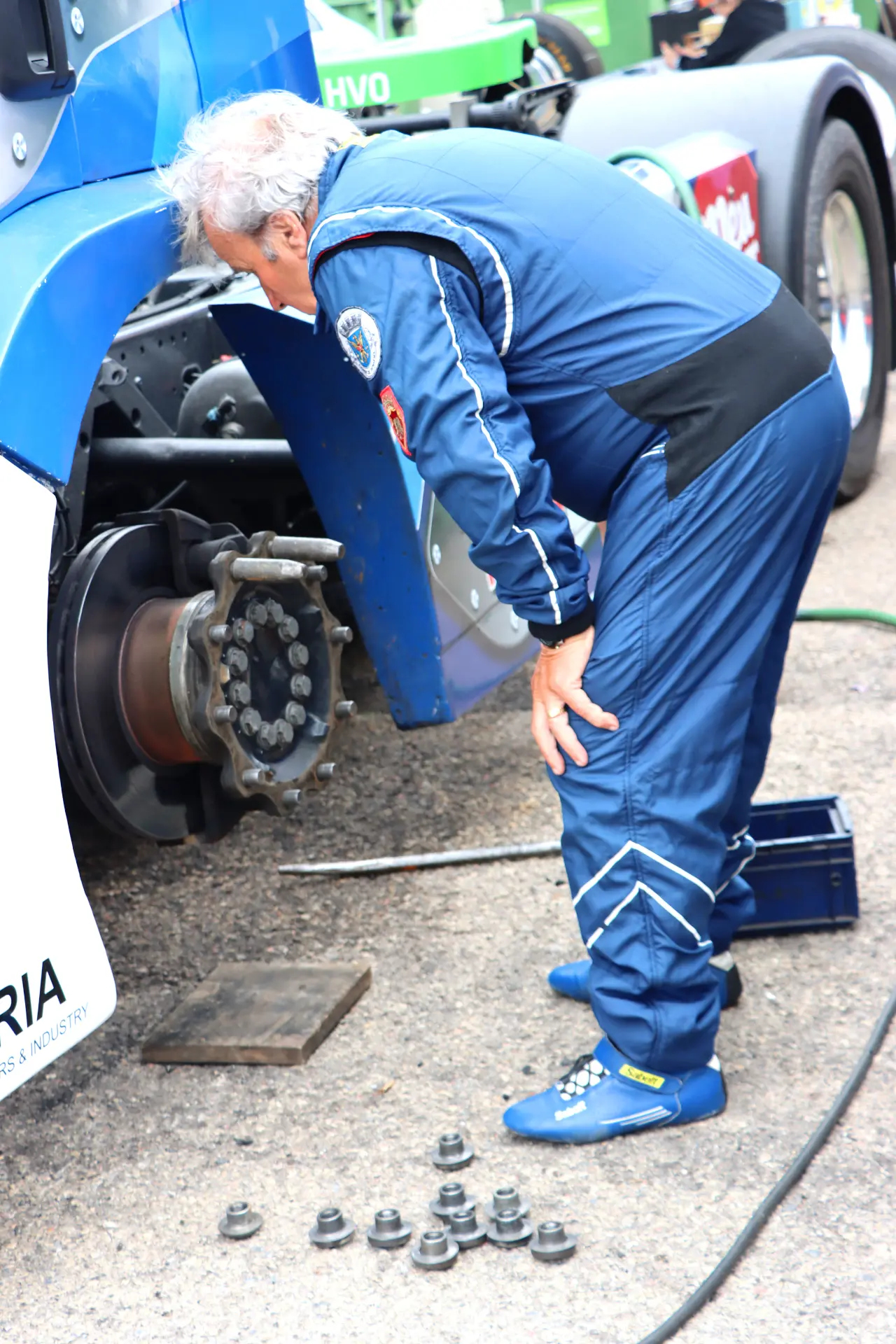 Un hombre con traje de carrera azul inspecciona el cubo de la rueda expuesto de un camión en el Valencia NASCAR Fest 2025; hay pernos y herramientas en el suelo, capturando la esencia de las carreras de camiones España y camiones americanos tuneados.