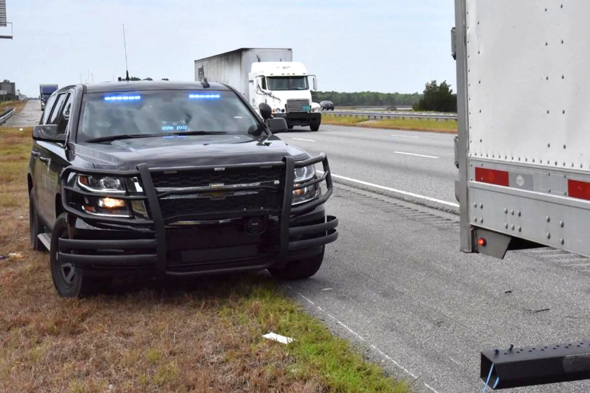 Una camioneta policial con luces azules intermitentes está estacionada detrás de un camión blanco al costado de una carretera, posiblemente realizando inspecciones en la carretera o controles a camiones.