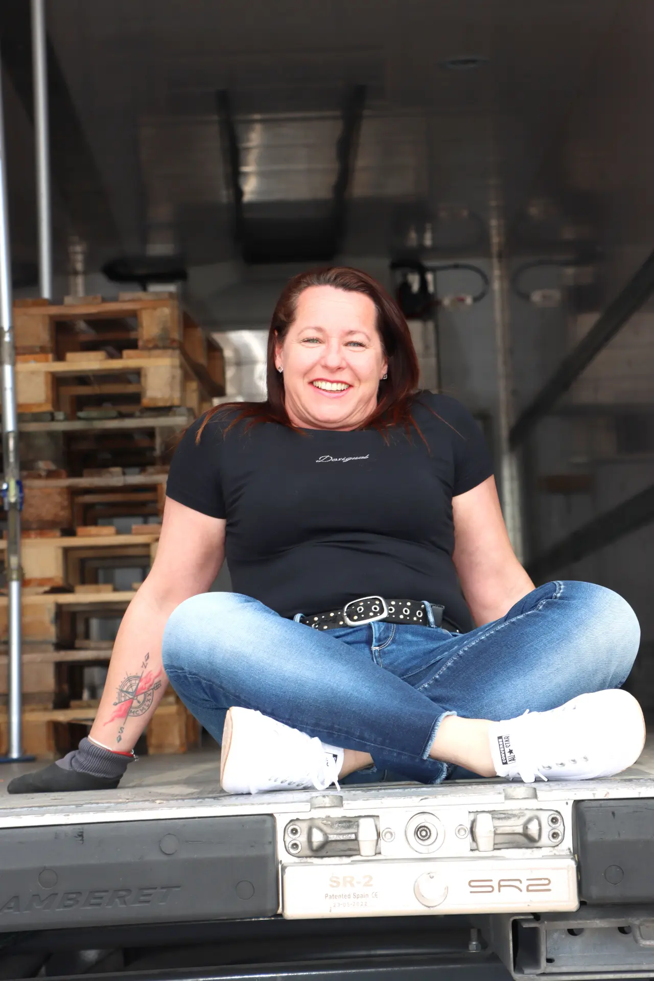 Una mujer sonriente con jeans se sienta con las piernas cruzadas en el cajón de un camión, con paletas de madera apiladas a su lado, sosteniendo un cuaderno con borrador automático.