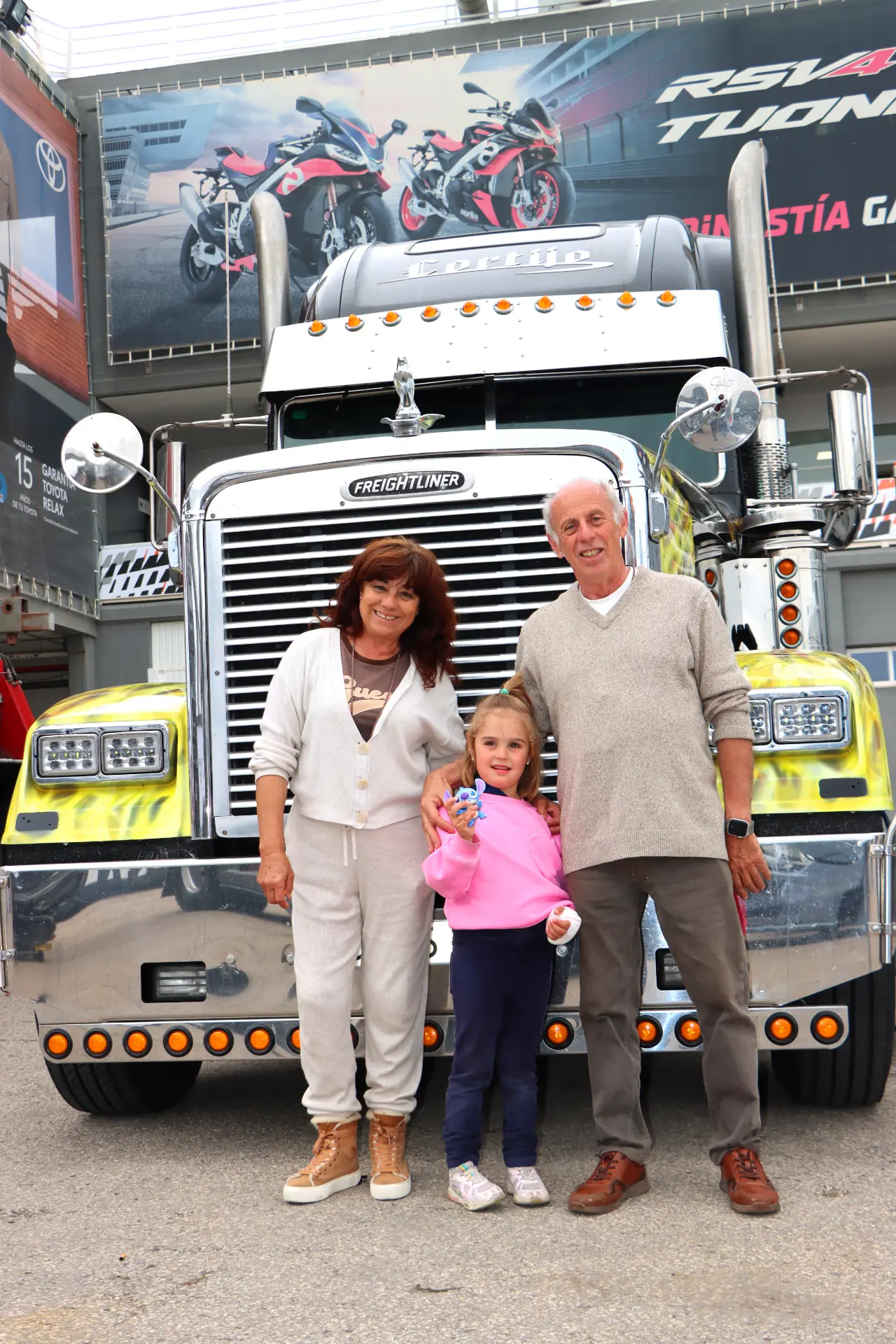 Una familia sonriente de tres personas se encuentra frente a un Freightliner amarillo brillante en el Valencia Nascar Fest 2025, celebrando los camiones americanos tuneados en este animado evento al aire libre.