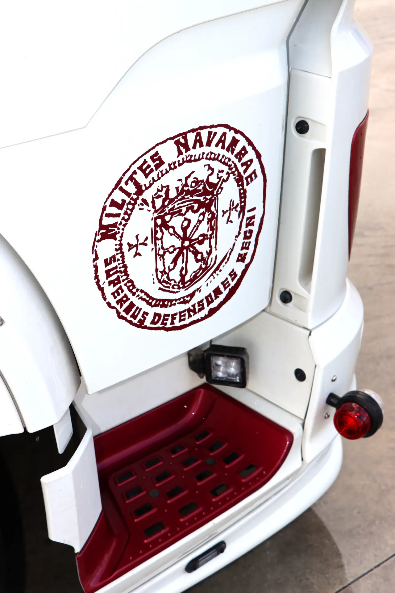 Un vehículo blanco con un emblema marrón y un texto en latín que dice "Milites Navarræ" en su costado sobre un escalón rojo.