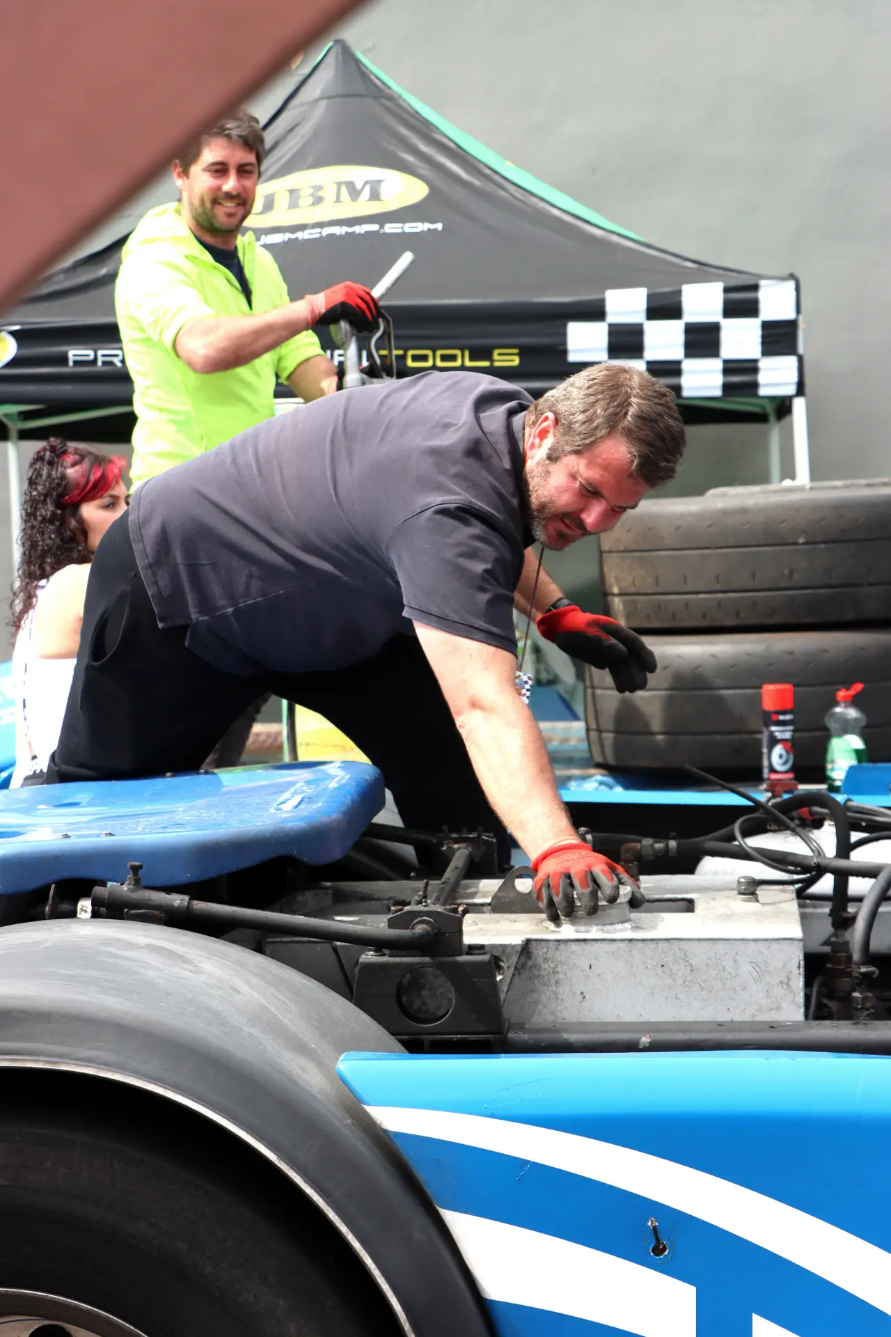 Dos hombres trabajan en un coche de carreras azul; uno está inclinado sobre el motor, mientras que el otro está detrás sonriendo en el Valencia NASCAR Fest 2025, donde las carreras camiones España y camiones americanos tuneados también son puntos destacados.