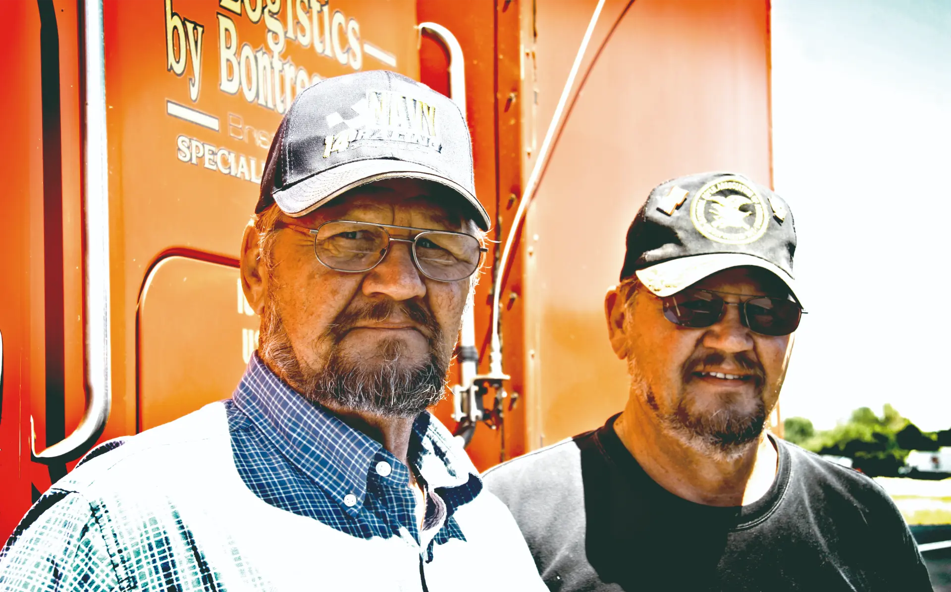 Dos hombres mayores con gorras y gafas están parados frente a un camión naranja brillante en un día soleado, posiblemente después de inspecciones en la carretera.