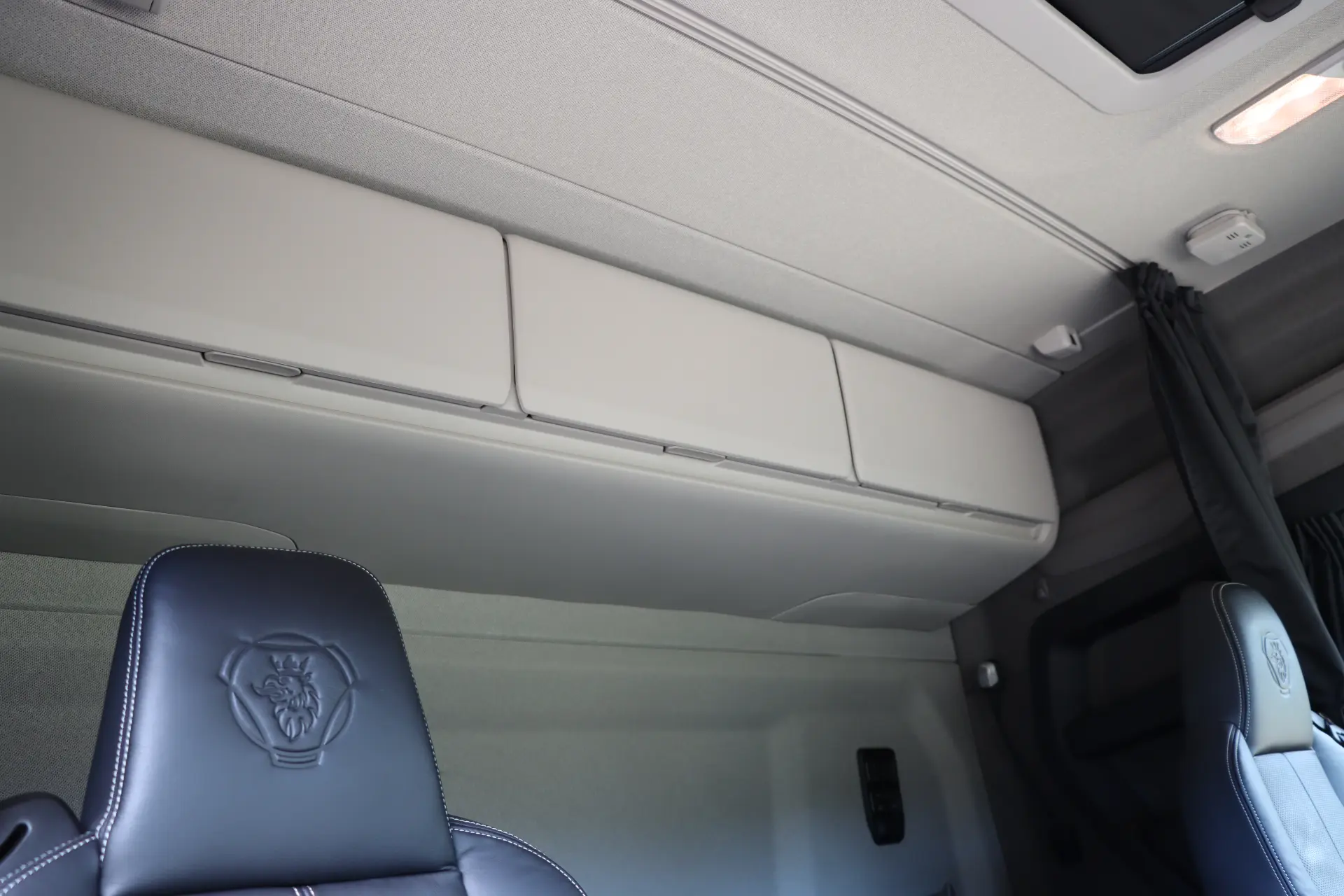 Compartimentos de almacenamiento superiores dentro de la cabina de un vehículo sobre un asiento negro con un logotipo en relieve, presentados en el Scania 40R eléctrico durante nuestra prueba en ciudad y carretera.