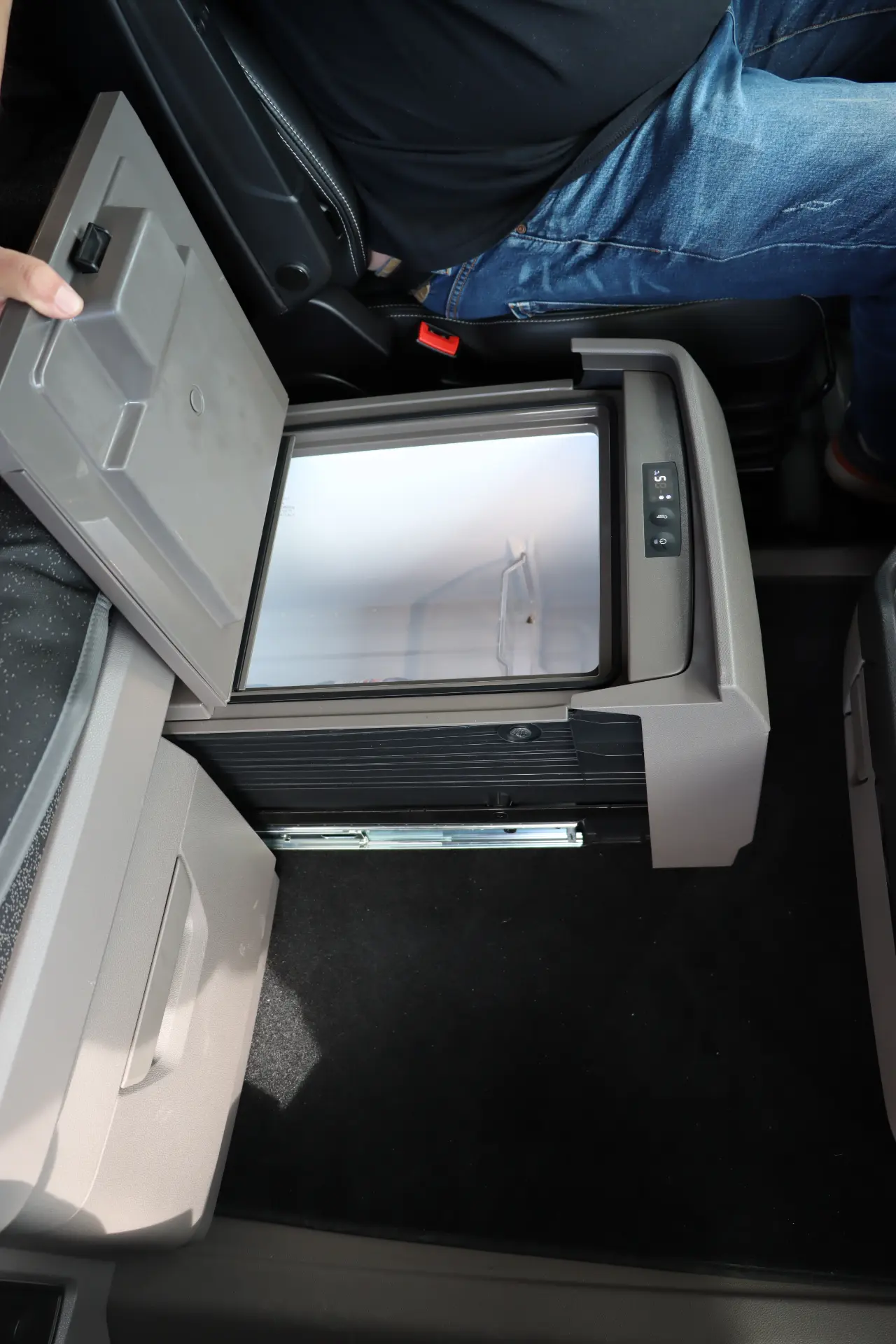 Una persona abre un mini refrigerador entre los asientos de un automóvil estacionado en la carretera, revelando un espacio interior vacío.