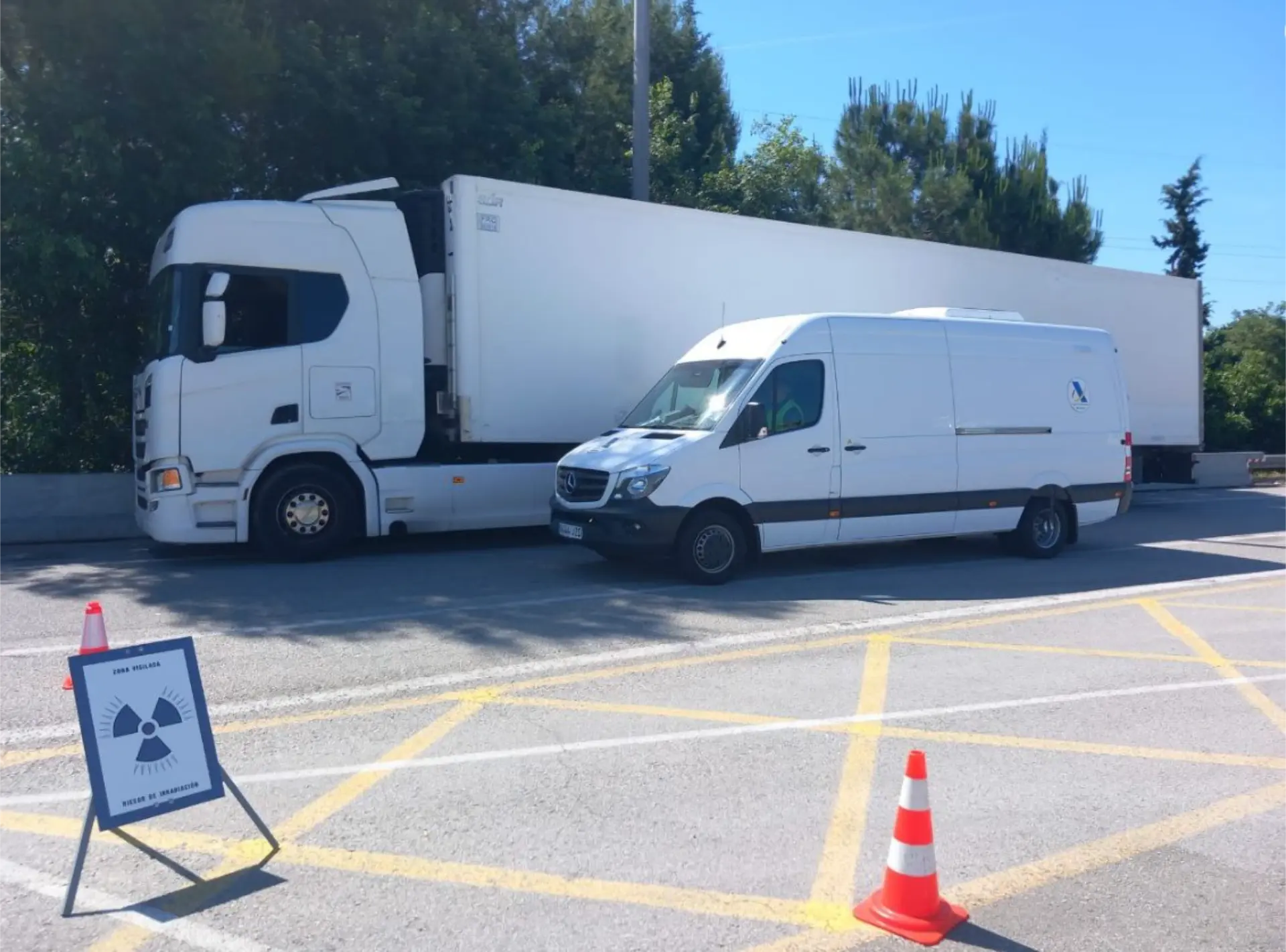 Un camión y una furgoneta con una señal de advertencia de radiación están estacionados cerca de conos de carretera y una señal de precaución durante una operación policial de macrocontrol AP-7.